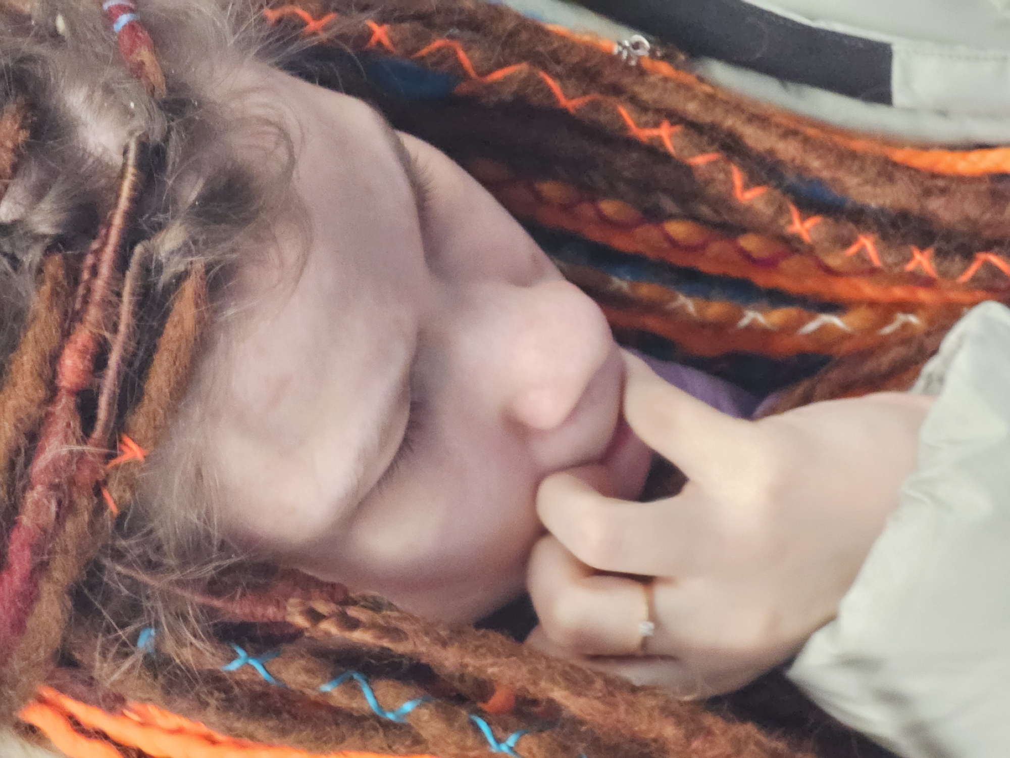 A person lies with their face sideways and eyes closed. Their hand touches their face. They wear a ring on their ring finger. The person has brown dreadlocks with colorful wraps. The environment is soft and blurry, possibly a bed or couch. The location and forensic details are unavailable from the image.
