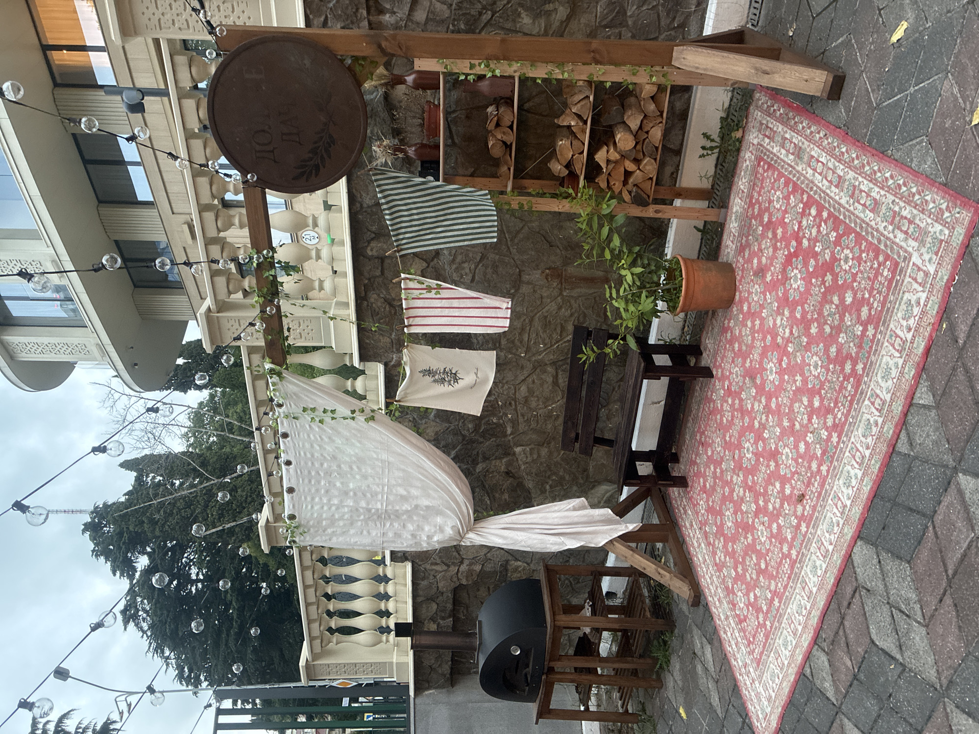 A rustic outdoor seating area is set up in front of a stone wall, likely on a patio or courtyard, in Sochi, Russia. The scene is adorned with string lights featuring exposed bulbs, creating a festive atmosphere. A black pizza oven sits on a wooden stand to the left, while a dark wooden bench is positioned centrally. A large, ornate red and white rug covers the patterned brick ground. To the right, a wooden shelf unit displays firewood and decorative vases. Several pieces of fabric, possibly towels or banners, hang on a clothesline. A potted green plant adds a touch of nature to the arrangement. In the background, a modern building with multiple stories and ornate balconies suggests an urban or resort setting. The overall impression is one of a cozy and inviting space, perhaps for outdoor dining or relaxation.