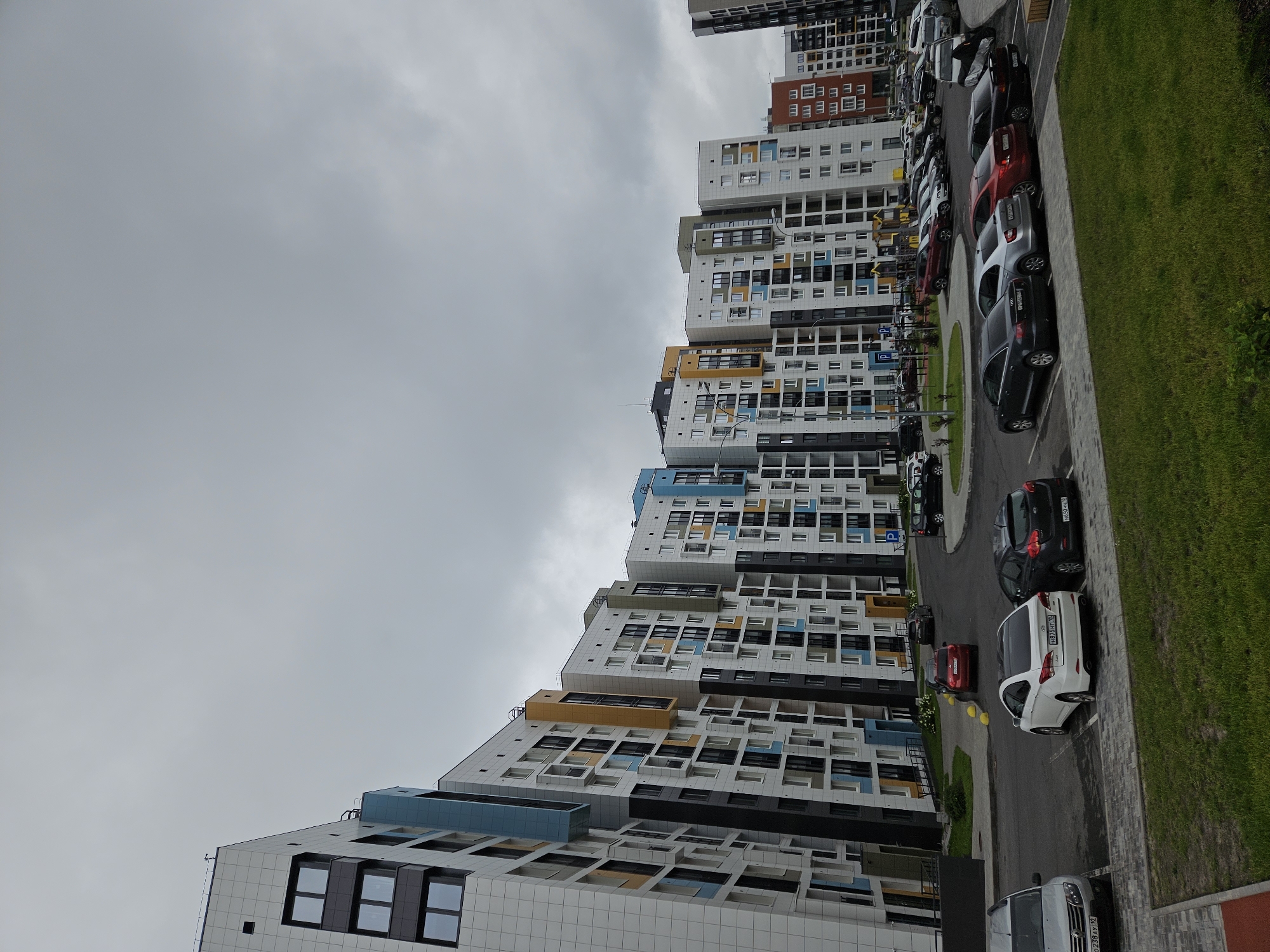 An overcast, wide-angle, eye-level shot captures a row of modern, colorful apartment buildings in a city. The buildings are characterized by their white facades accented with blocks of bright colors such as blue, yellow, and orange, and feature numerous windows. In the foreground, a parking lot is filled with various cars, including a white sedan, a dark SUV, and a silver hatchback. The ground is a mix of asphalt and paved walkways, with patches of green grass. Beyond the immediate parking area, more cars are visible, and to the right, a section of another building, this one red with white windows, is present. The sky is a uniform gray, suggesting a cloudy day.
