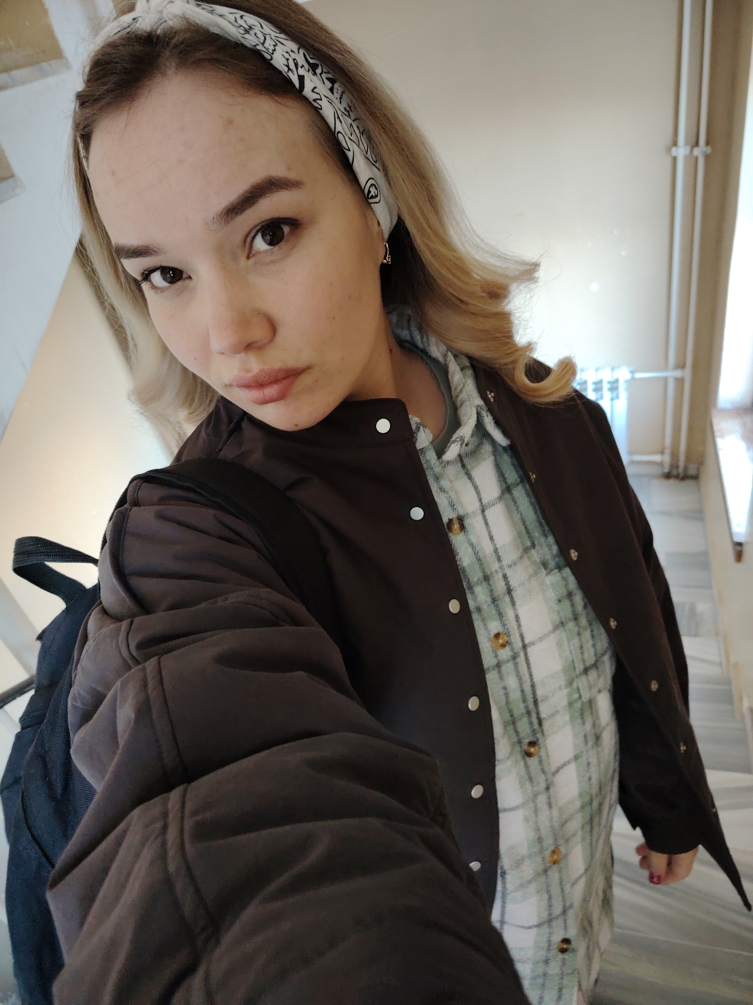 A young woman takes a selfie while standing on a marble staircase. She has fair skin, light brown eyebrows, and dark eyes, and her blonde hair is styled with a curl and a bandana. She wears a light green and white plaid shirt under a brown jacket, with a black backpack over her left shoulder. The environment includes light beige walls, a white radiator, and a window with vertical bars. The location is Марковский, Russia.