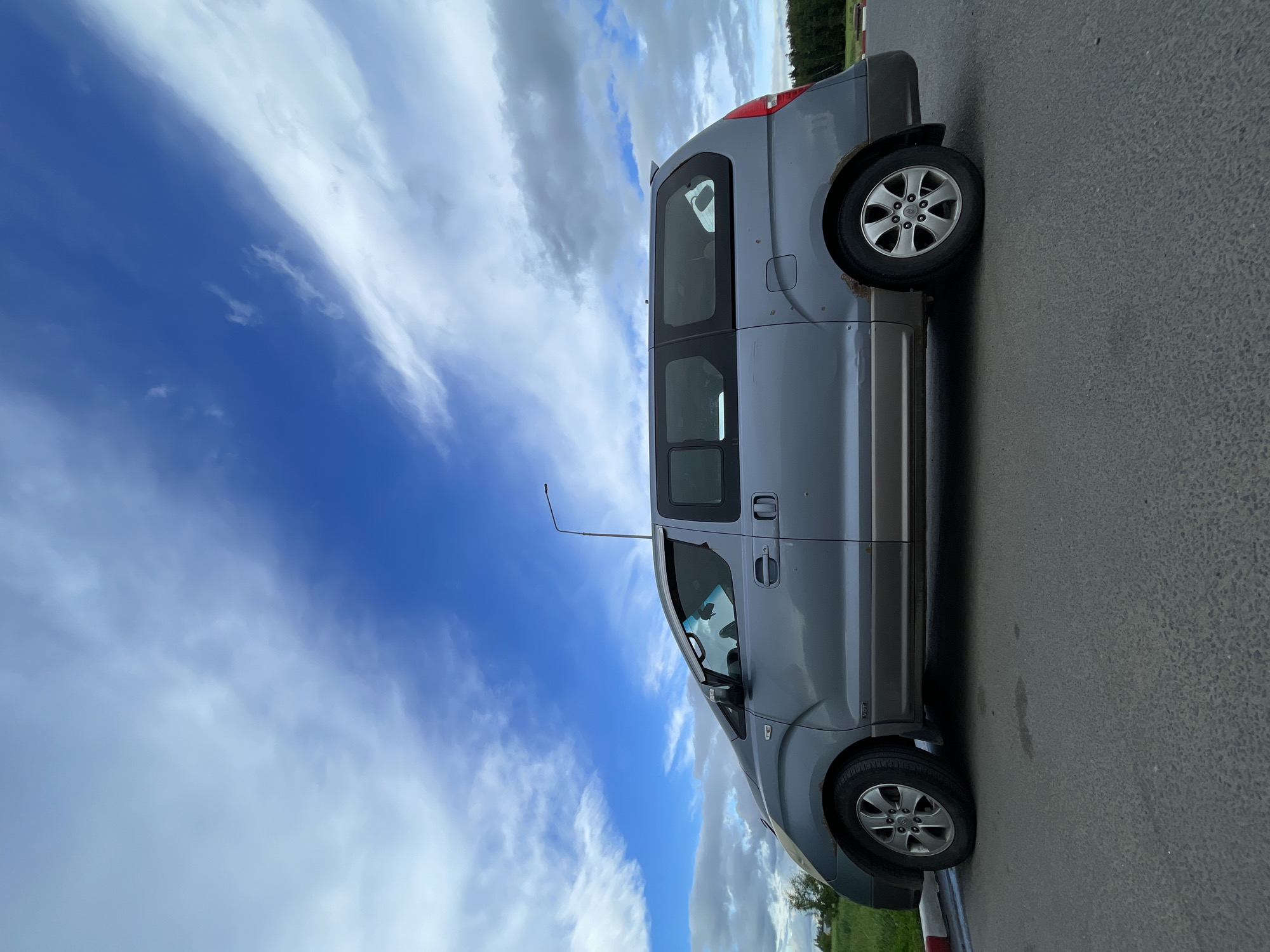 A gray Hyundai Starex minivan is parked on the side of an asphalt road in Бавыкино, Russia. A lamppost stands next to the vehicle. The sky behind the minivan is partly cloudy with blue patches visible. The area behind the van appears to be a grassy field with some trees in the background. A red and white striped curb is visible along the edge of the road.