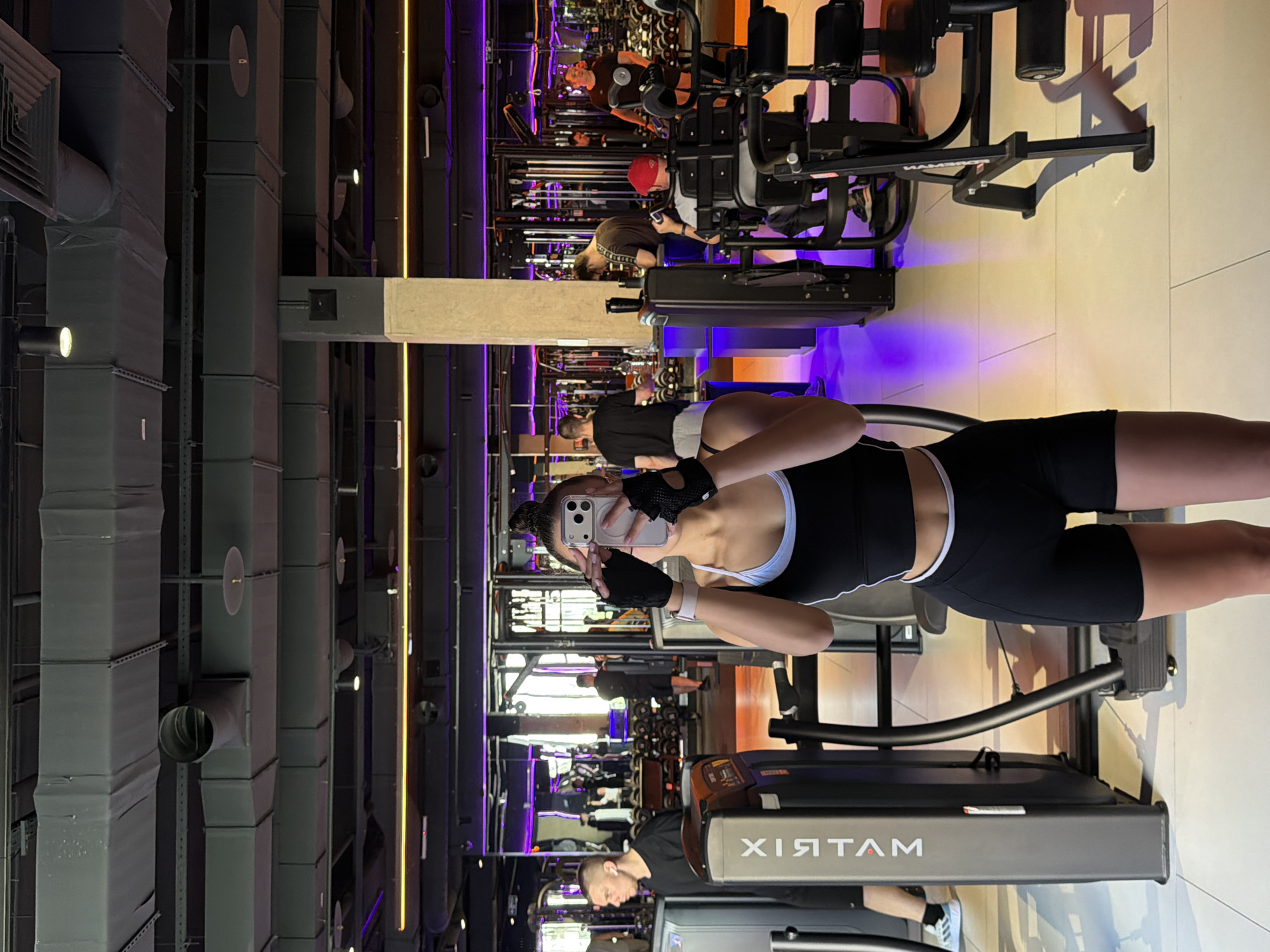 A woman in a black and white workout outfit is taking a selfie in a gym, using a white smartphone held in her left hand. She has a messy bun hairstyle and is wearing black fingerless gloves. In the background, other gym-goers are using various exercise machines. Ductwork and lighting fixtures are visible on the ceiling. The treadmill she is standing on has the word "MATRIX" on it. The exact location in Podolsk, Russia cannot be determined from the image.