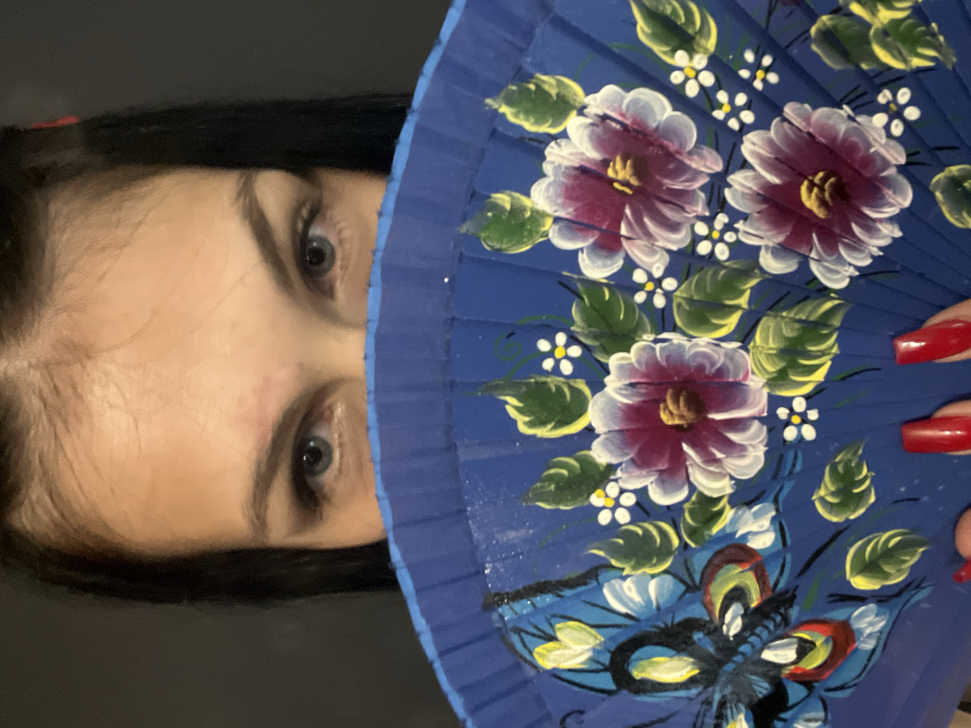 The media file features a close-up of a person's upper face and a hand holding a decorative fan, set against a dark, indistinct background. The person's head is horizontally oriented within the portrait-mode image, with their dark hair visible along the top edge and forehead/eyes occupying the left side of the frame. The eyes are light-colored and look directly at the viewer.
The main object is a blue hand fan, which covers most of the lower and right side of the person's face. The fan is adorned with vibrant painted designs, including large pink or maroon flowers with white outer petals and yellow centers, surrounded by green and yellow leaves. Smaller white flowers with yellow centers are scattered throughout the design. Towards the bottom of the fan, a multi-colored butterfly in shades of blue, black, yellow, and red is visible. The person's hand, holding the fan, shows fingernails painted with bright red polish.
The activity depicted is a person posing for a photograph, partially concealing their face with the ornate fan. The direct gaze suggests an intentional and expressive pose.
Notable details include the bold colors of the fan's design and the contrast with the dark background. The lighting highlights the fan and the person's eyes. There is no visible text in the image. The setting is ambiguous, likely indoors or in a controlled environment due to the plain, dark background.