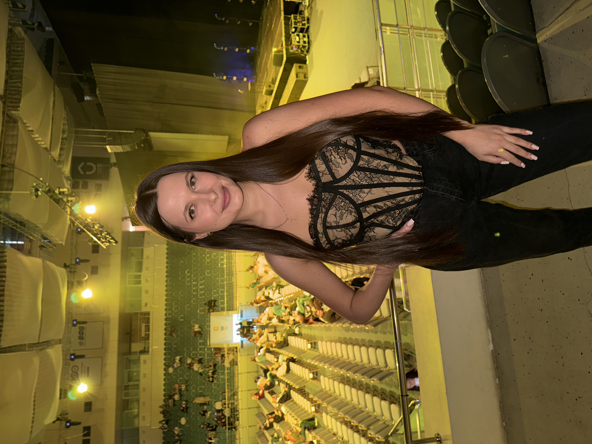 A young woman is captured in an indoor venue, likely an arena or concert hall, from an elevated perspective. The image is rotated 90 degrees clockwise, showing her standing upright and facing the viewer with a gentle smile. She has long, straight dark brown hair that cascades over her shoulders and chest. Her attire consists of a black, possibly strapless or spaghetti-strap top featuring intricate beige lace patterns, resembling a corset or bustier, paired with dark-colored pants or jeans. A delicate silver-toned necklace with a small cross pendant is visible around her neck, and her fingernails are painted with light-colored polish.
The background reveals numerous rows of green-cushioned seats with gold-toned frames, extending into the distance. Many of these seats are occupied by a crowd of people, suggesting a live event. In the far background, a stage area is discernible, equipped with performance lighting, including blue and white lights, and audio equipment such as speakers and rigging. The venue is illuminated by artificial, warm yellow lights, indicating an evening or nighttime setting.
Visible text includes a sign that appears to read "B&B" or "BOB", another sign with a logo resembling "C>", and a more distant sign that might say "UNI" or "UNO". The overall scene evokes the lively atmosphere of a concert or a similar public performance.
