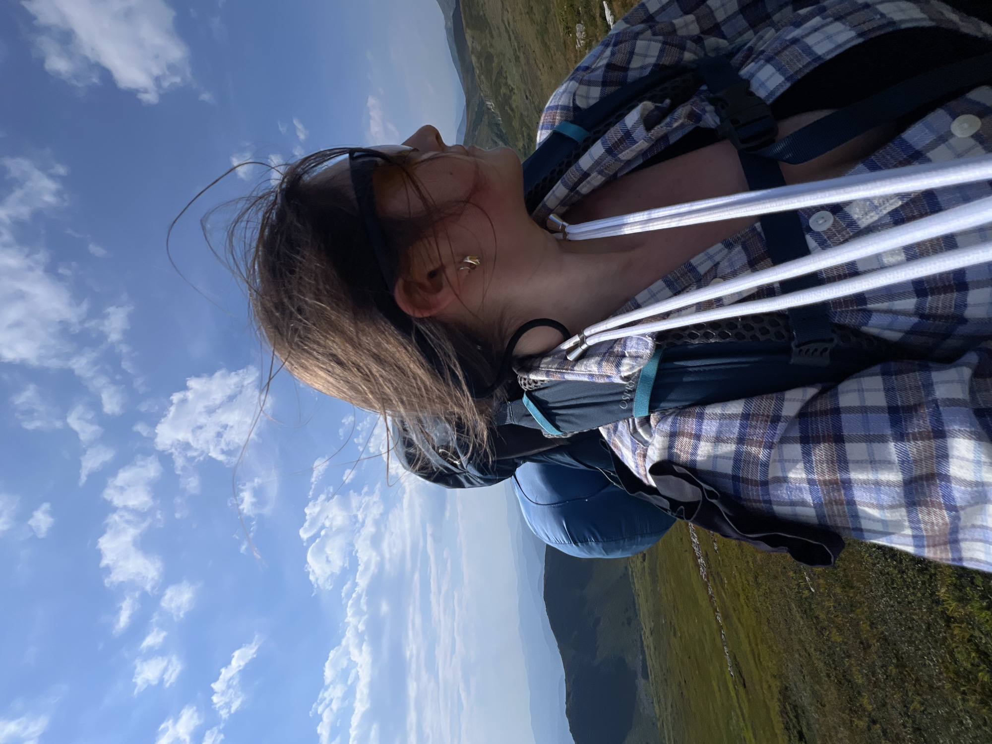 A woman is captured in a close-up, selfie-style shot while hiking in the mountains. She is wearing a blue and white plaid shirt over a dark-colored bra and sunglasses. A large blue backpack is on her shoulders, with white straps visible in front of her. Her hair is windswept, blowing across her face. The background shows rolling green hills and mountains under a bright blue sky with scattered white clouds. The location is the Carpathian Mountains, near the village of Luhy, Ukraine, known for its scenic hiking trails and panoramic vistas. The overall impression is one of adventure and natural beauty.