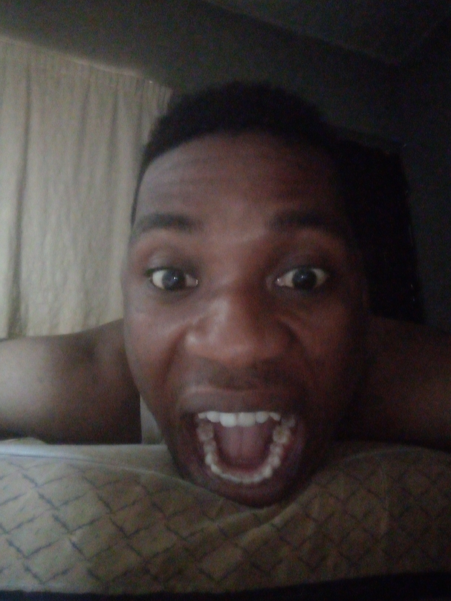 A close-up, eye-level shot shows a man with dark skin lying on a patterned surface. He is facing the camera with wide eyes and an open mouth, revealing his teeth. Part of his shoulder is visible on the left side. Behind him, a light-colored curtain hangs on the left, and a dark area is on the right. The location is Unknown, Unknown.