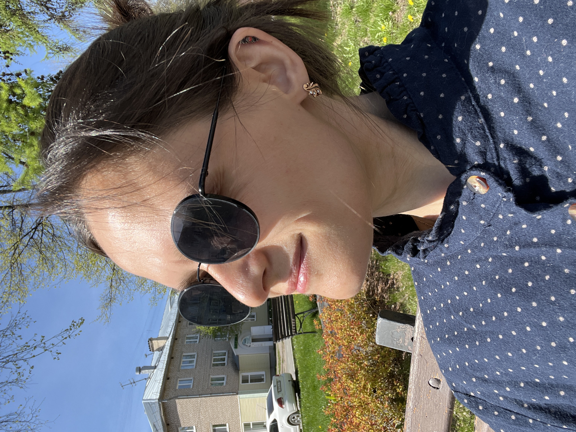 A woman with dark hair in a messy bun is wearing round sunglasses and a navy blue shirt with white polka dots and ruffles at the neckline, sitting on a wooden bench. The woman has a piercing on her ear. Behind her is a park with a light green building, a white car, and a black park bench. The location is likely Chaikovsky, Russia.