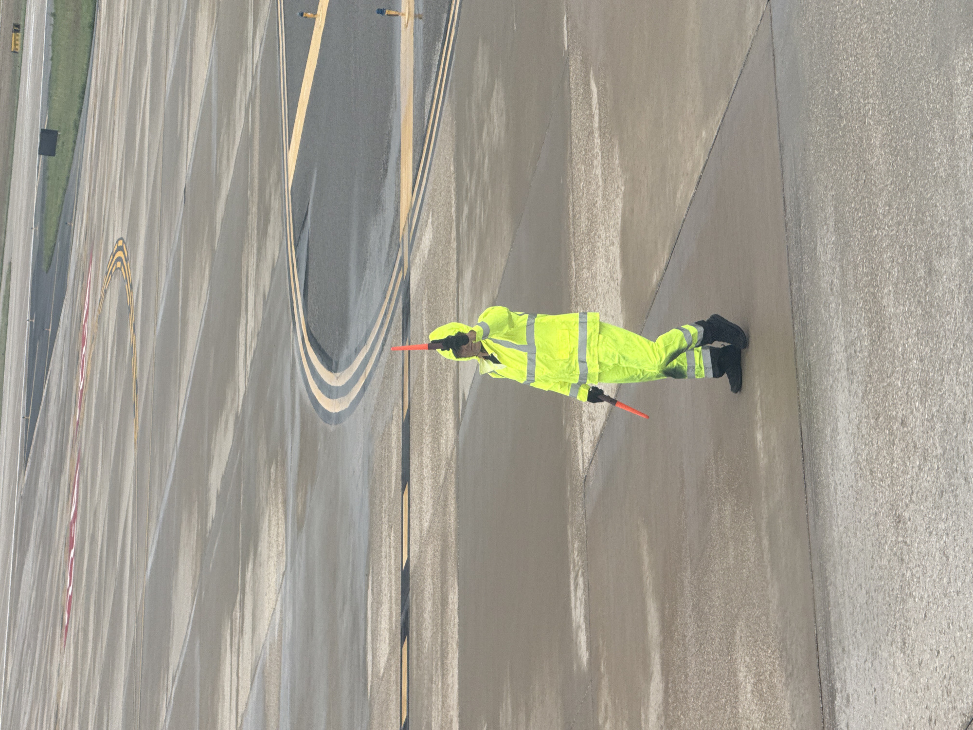 Aviation traffic controller at San Antonio International Airport in San Antonio, Texas, signals an aircraft on the tarmac. The controller wears a neon yellow safety suit, black gloves, and boots. They hold orange wands in both hands. The tarmac shows yellow lines, markings, and a water-like surface.