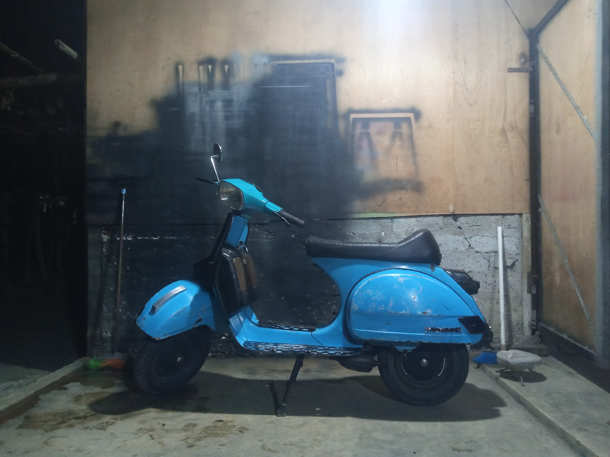 A blue Vespa scooter is parked in what appears to be a dimly lit garage or workshop with concrete floors and wooden walls. The scooter is positioned centrally in the frame, facing to the left. The background wall has a textured, possibly spray-painted, mural of abstract shapes and lines in dark colors. To the left of the scooter, a broom and a mop lean against a dark, shadowy wall. To the right of the scooter, a white pipe stands upright, and a light-colored object, possibly a bird feeder or a small statue, sits on the concrete. The scene is static and devoid of human presence. The overall impression is one of a storage or working space, possibly in a rural or industrial setting within Balee Rastong, Indonesia, given the context.