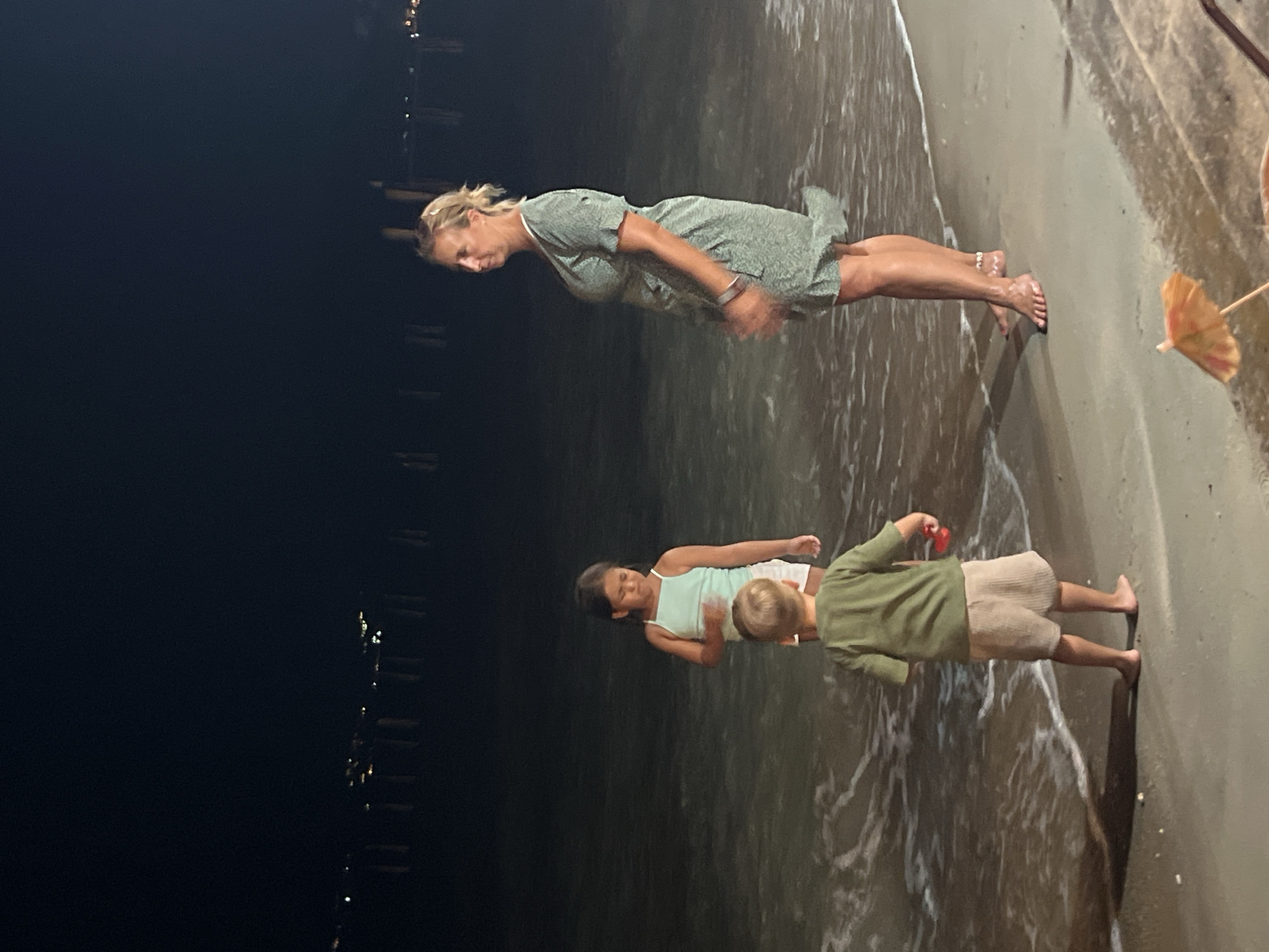 In an outdoor, nighttime scene at the beach in Unknown, Unknown, a woman and two young children stand near the water's edge. The woman, wearing a green patterned dress, faces the left side of the frame, her arms blurred by movement. A young girl in a light blue tank top and white shorts stands to the left of the woman, looking down at something in the water. A young boy, wearing a green t-shirt and beige shorts, stands in front of the girl, facing away from the camera. He holds a red toy in his right hand. The water laps at their feet, and the sand is wet. In the background, the dark ocean stretches out, with distant lights visible on the shore. A small, orange umbrella is visible in the lower right corner of the frame, suggesting a nearby seating area.