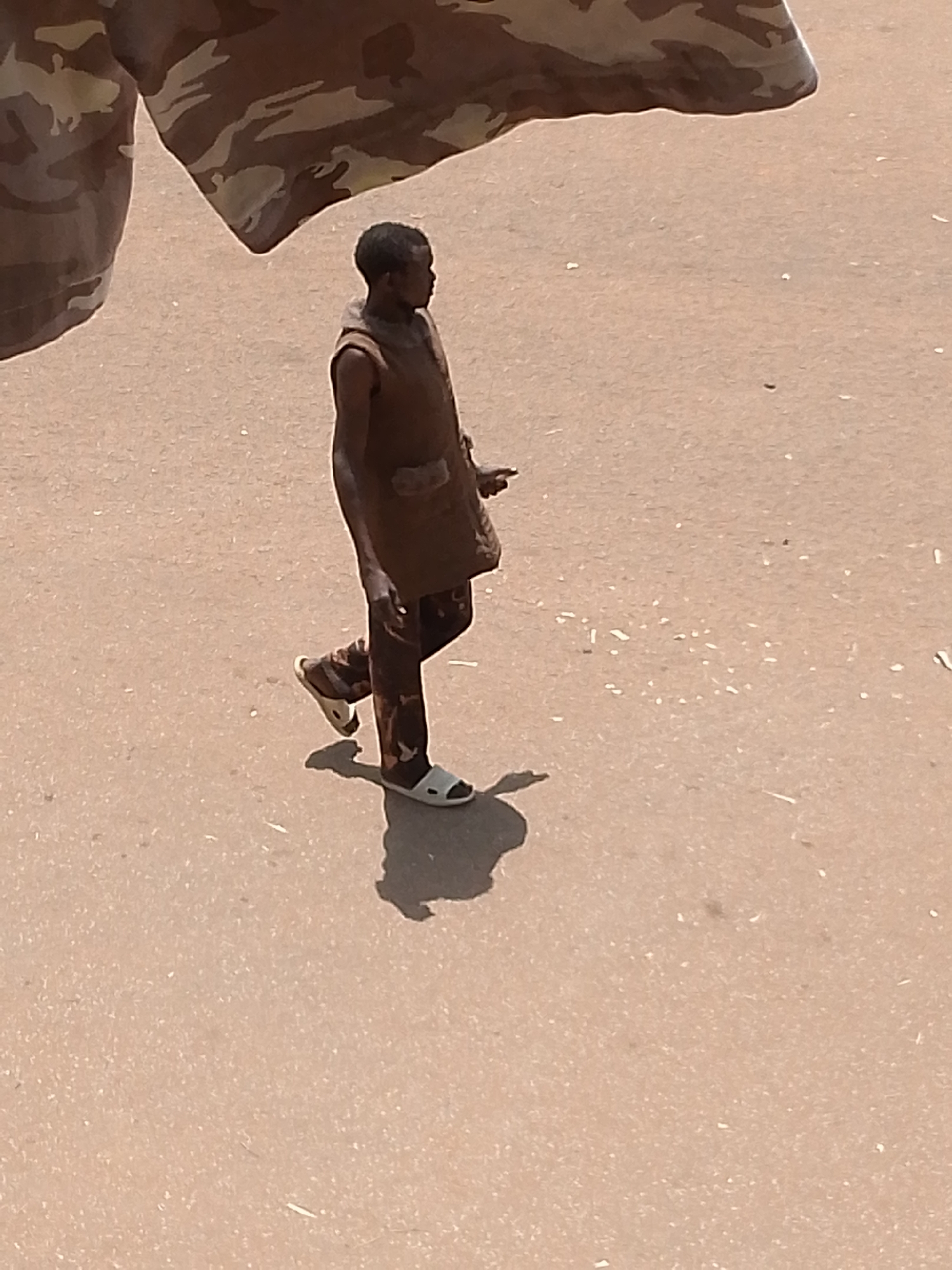 A person is walking on a sandy surface in Bukuru, Nigeria. The person is wearing a brown sleeveless vest over a patterned shirt and pants, and white sandals. The person's left leg is extended forward, and the right leg is bent at the knee, indicating a walking motion. A shadow of the person is visible on the ground behind them. In the upper portion of the image, a large piece of camouflage fabric is draped, obscuring the top of the scene and casting a shadow. The ground appears to be dusty or sandy, with small white specks scattered across its surface.