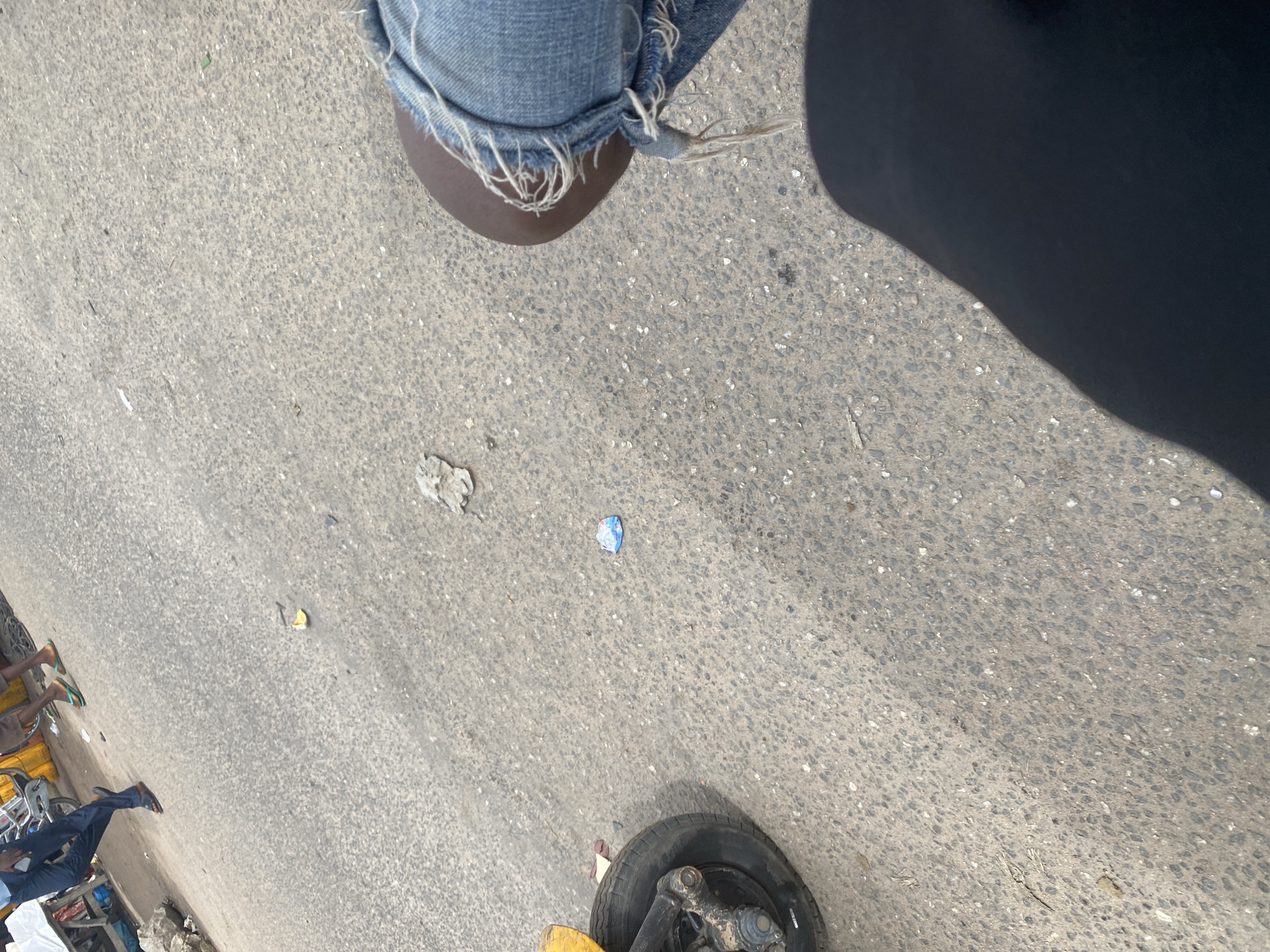 The image shows a ground-level view of a street, likely in Jalingo, Nigeria. The surface is asphalt, textured with small stones. In the upper left, a person's legs are visible, wearing distressed denim jeans with frayed hems and dark brown shoes. To the right of the legs, a large shadow is cast, suggesting bright sunlight. Scattered on the ground are pieces of litter, including a crumpled white paper and a small blue and white wrapper. Further down the street, more people are visible. One person is wearing blue pants and dark sandals, with their legs crossed. Another person is wearing flip-flops. There are glimpses of market stalls or carts, suggesting a commercial area. A yellow object and a black tire with its mechanical components are also visible in the lower portion of the frame, possibly part of a vehicle or cart. The scene appears to be outdoors during the daytime, under sunny weather conditions, typical of a busy street environment.