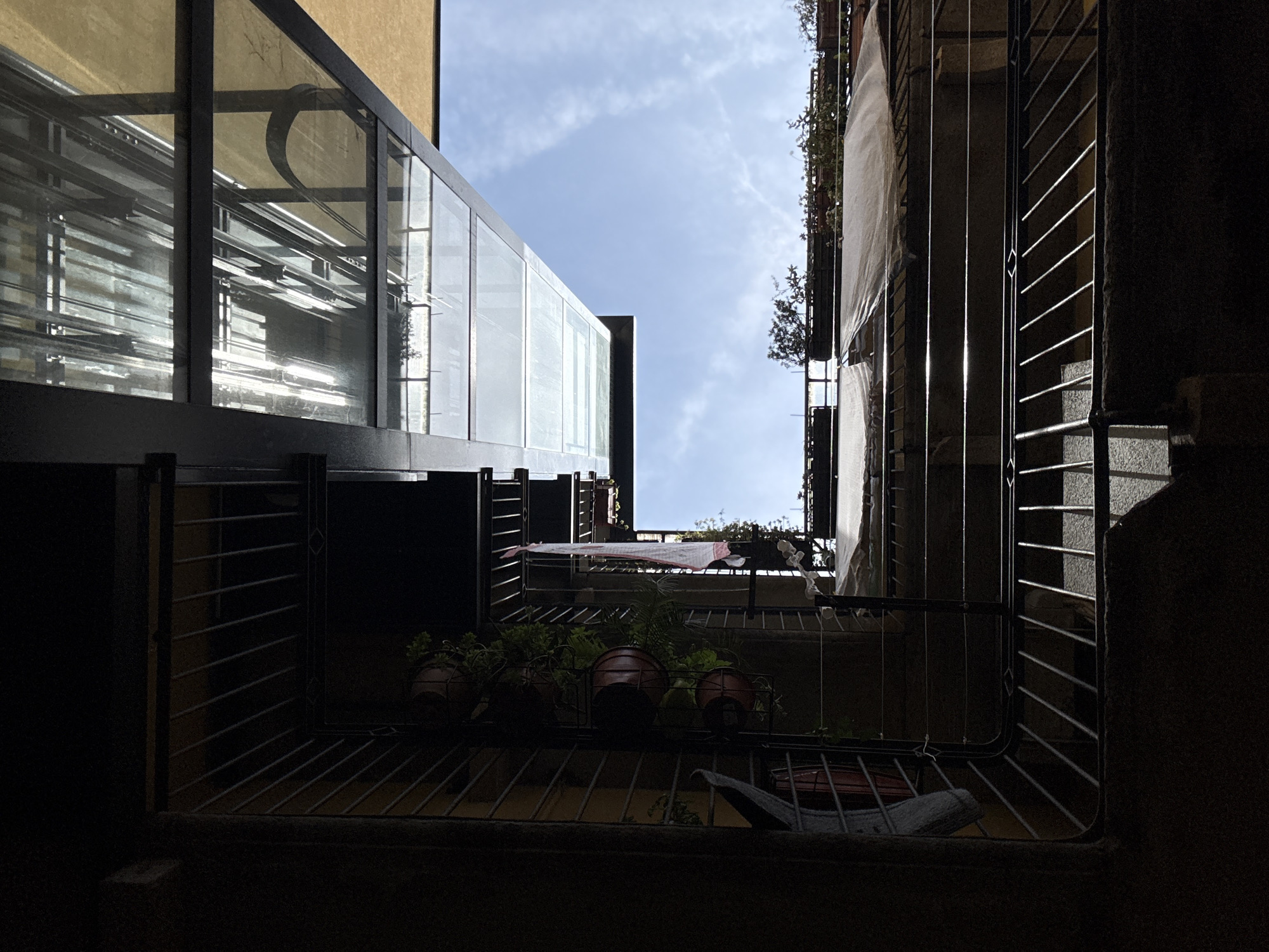 The image depicts a view looking upwards from a narrow courtyard in Milan, Italy. The architecture suggests a multi-story building with balconies and metal railings. Several potted plants are visible on a rack attached to one of the balconies, with one white and pink garment hanging on a clothesline. Above, a modern glass-covered structure with metal beams is visible, reflecting the sky. The sky is blue with scattered clouds. The lighting suggests it is daytime, with strong shadows cast by the buildings. The overall scene appears to be an urban environment characterized by functional, somewhat utilitarian architecture.
