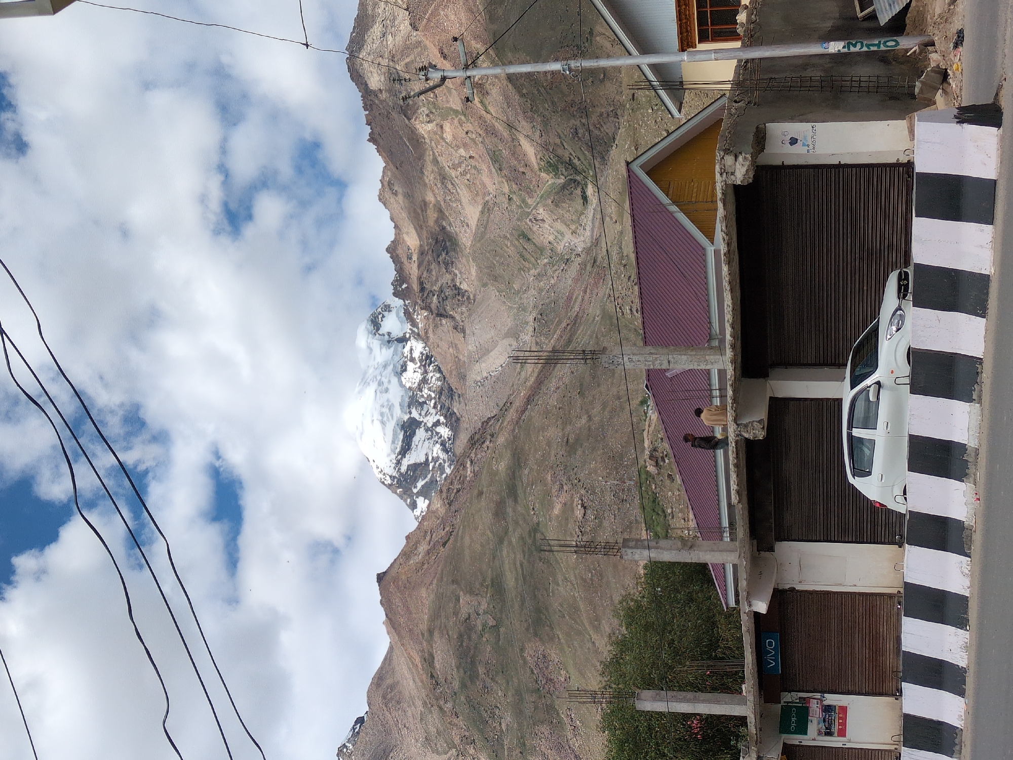 The image shows a street scene in Padum, India, with a snow-capped mountain range in the background. In the foreground, there are several shops with rolled-down shutters, a white car parked on the street, and a concrete barrier. Two men are standing on the roof of one of the shops, observing the surroundings. Power lines are strung across the sky, and a utility pole is visible on the right side of the image. The buildings appear to be constructed with concrete and metal roofing. The mountain slopes are rocky and barren, with patches of green vegetation. The sky is partly cloudy, with patches of blue visible. The overall impression is a blend of urban infrastructure and natural landscape.