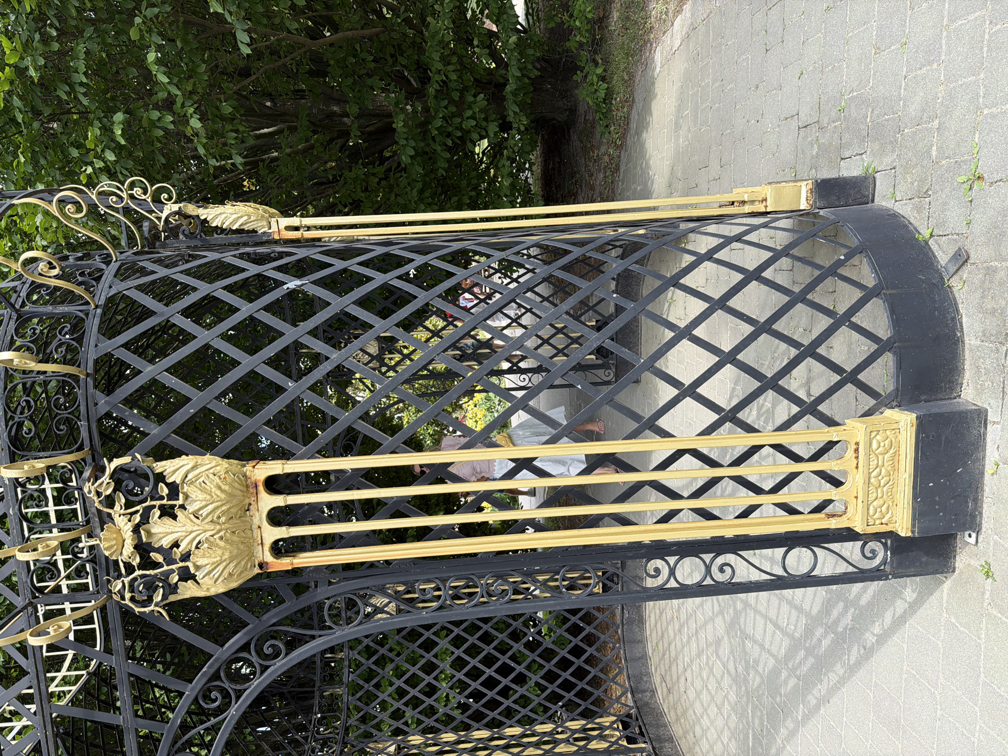 The image shows an ornate black and gold gazebo structure in a park. The gazebo has a diamond-patterned lattice design and is accented with decorative gold elements, including fluted columns topped with leafy motifs. Through the open entrance of the gazebo, two individuals are visible, one person is wearing a pink top and a light-colored skirt, carrying a yellow bag. Another person is partially visible behind them. The ground inside and outside the gazebo is paved with light-colored square stones. Lush green trees and foliage form the background. The scene appears to be set in a well-maintained public outdoor space. This is likely the Prater Park in Vienna, Austria, given the style of the gazebo and the overall park environment.