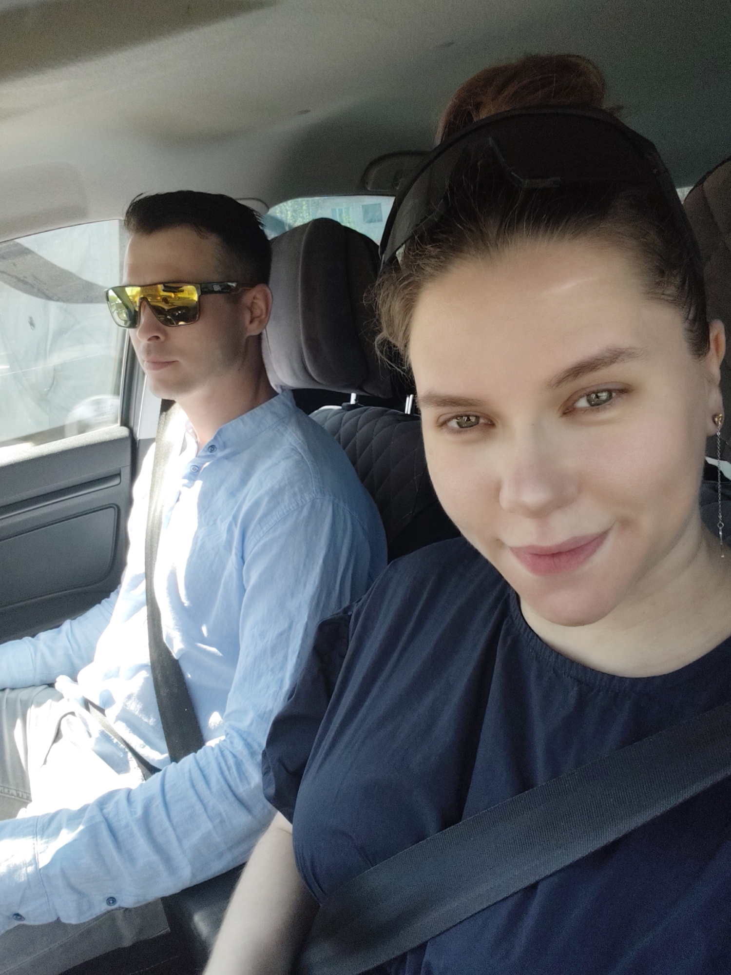 A selfie shows two people inside a car. A woman is in the passenger seat, angled toward the camera, wearing a dark blue shirt and a seatbelt, with dark hair in a bun and sunglasses on her head. She is smiling slightly. A thin, dangling earring is visible on her left ear. To her left, the driver is wearing sunglasses, a light blue button-down shirt, and a seatbelt. His dark hair is closely cropped. The interior of the car is visible, including grey seats and a car door panel. Outside the car, there is a window. The location may be Chaikovsky, Russia.