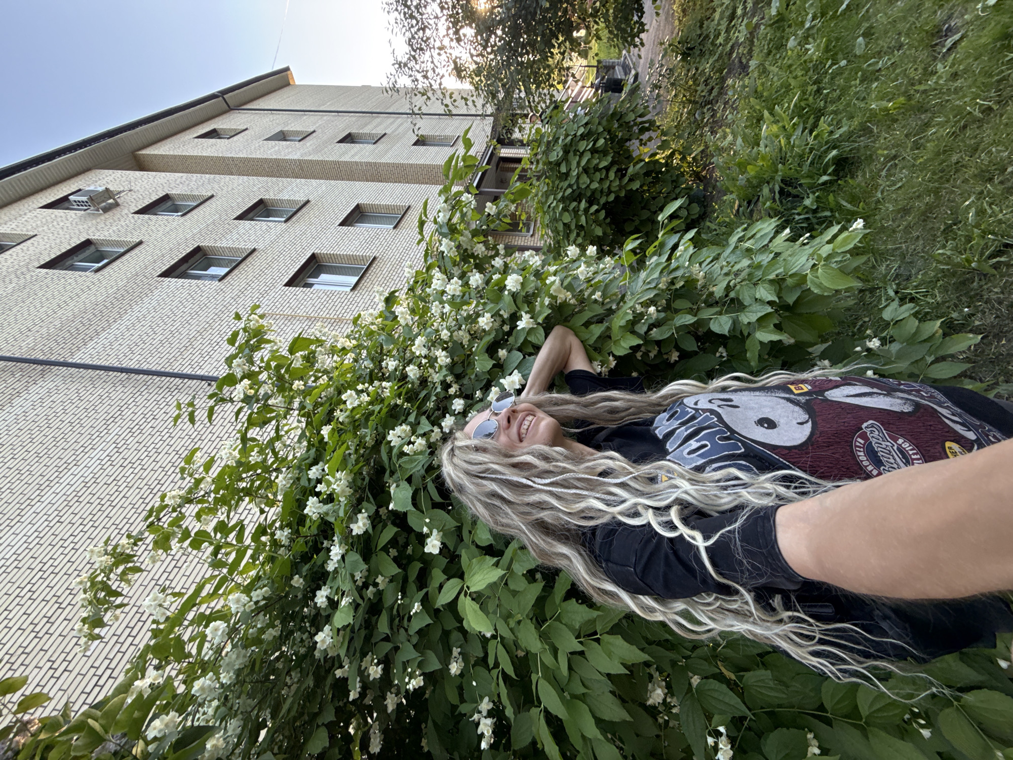 The image depicts an outdoor scene in Rusanovo, Russia, featuring a woman taking a selfie in front of a blooming shrub with a residential building in the background.
The central subject is a woman with very long, wavy, multi-toned blonde hair, including distinct white braids. She is wearing round, silver-rimmed sunglasses and a black t-shirt with a "Brooklyn Champs Athletics" graphic featuring a cartoon character (likely Snoopy). She is smiling, looking upwards and to her right, with her right hand behind her head. Her left arm is extended towards the camera, indicating she is taking a selfie.
She is positioned to the left of a large, dense shrub, heavily covered in small white flowers and dark green leaves, which dominates the left and central midground. Behind the shrub and to the right, a multi-story building constructed of light beige brick with dark mortar lines is visible. The building features multiple rectangular windows with dark frames, and a white exterior air conditioning unit is installed below one of the upper windows. A clear blue sky is seen above the building.
To the right of the main shrub, additional green foliage, smaller bushes, and trees are present, with sunlight illuminating parts of the leaves. The ground in the lower right consists of green grass and some darker patches of earth. A portion of a covered entrance or porch with steps is faintly visible on the right side of the building behind the foliage.