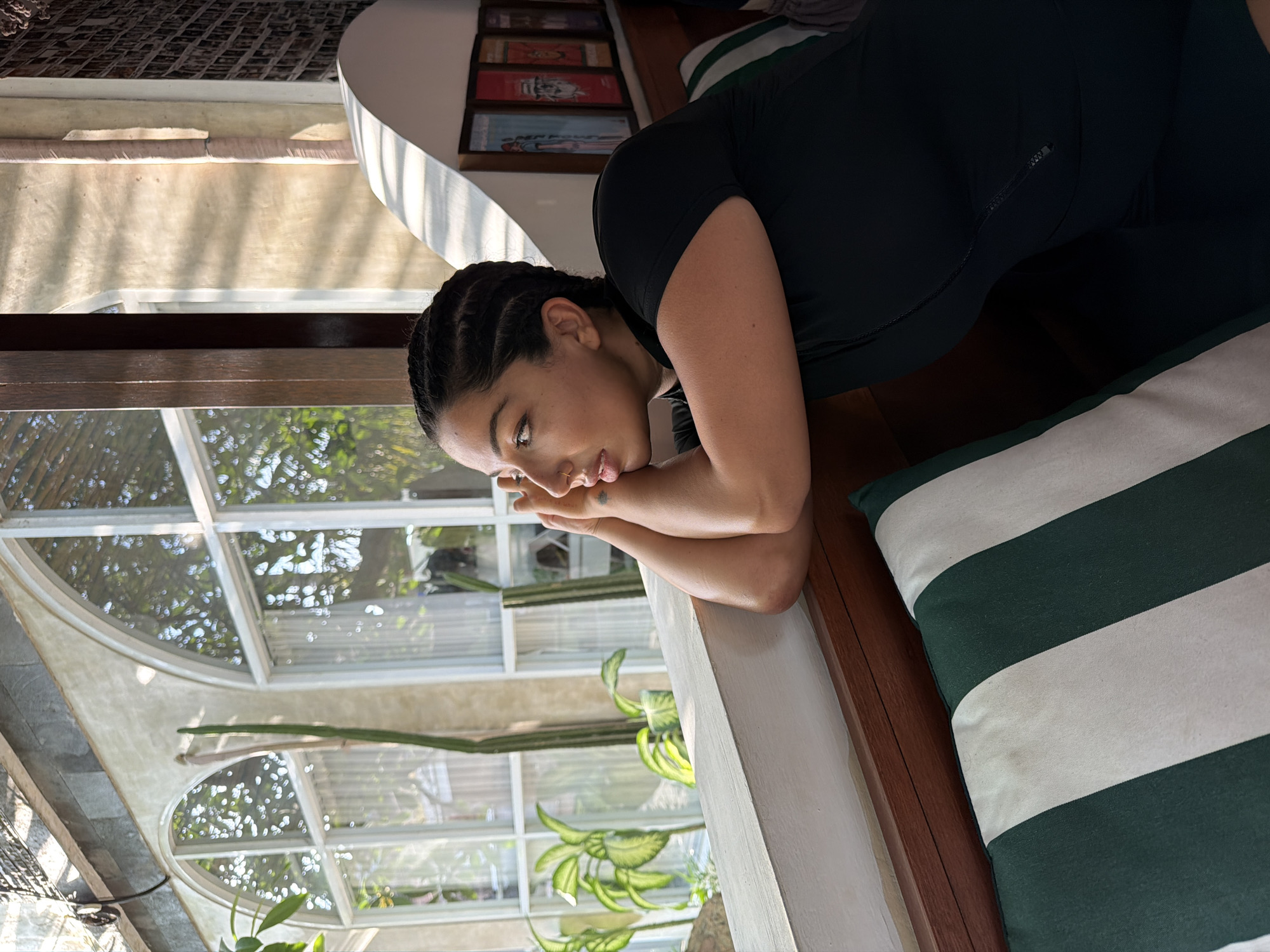 In the city of Mengwi, Indonesia, a woman with braided hair is leaning on a white ledge, looking out of a window. She is wearing a black short-sleeved shirt and is resting her head on her hands. Through the arched window, green plants and trees are visible, along with white sheer curtains. The room has a textured wall with shadows cast by sunlight. A striped green and white cushion is in the foreground.