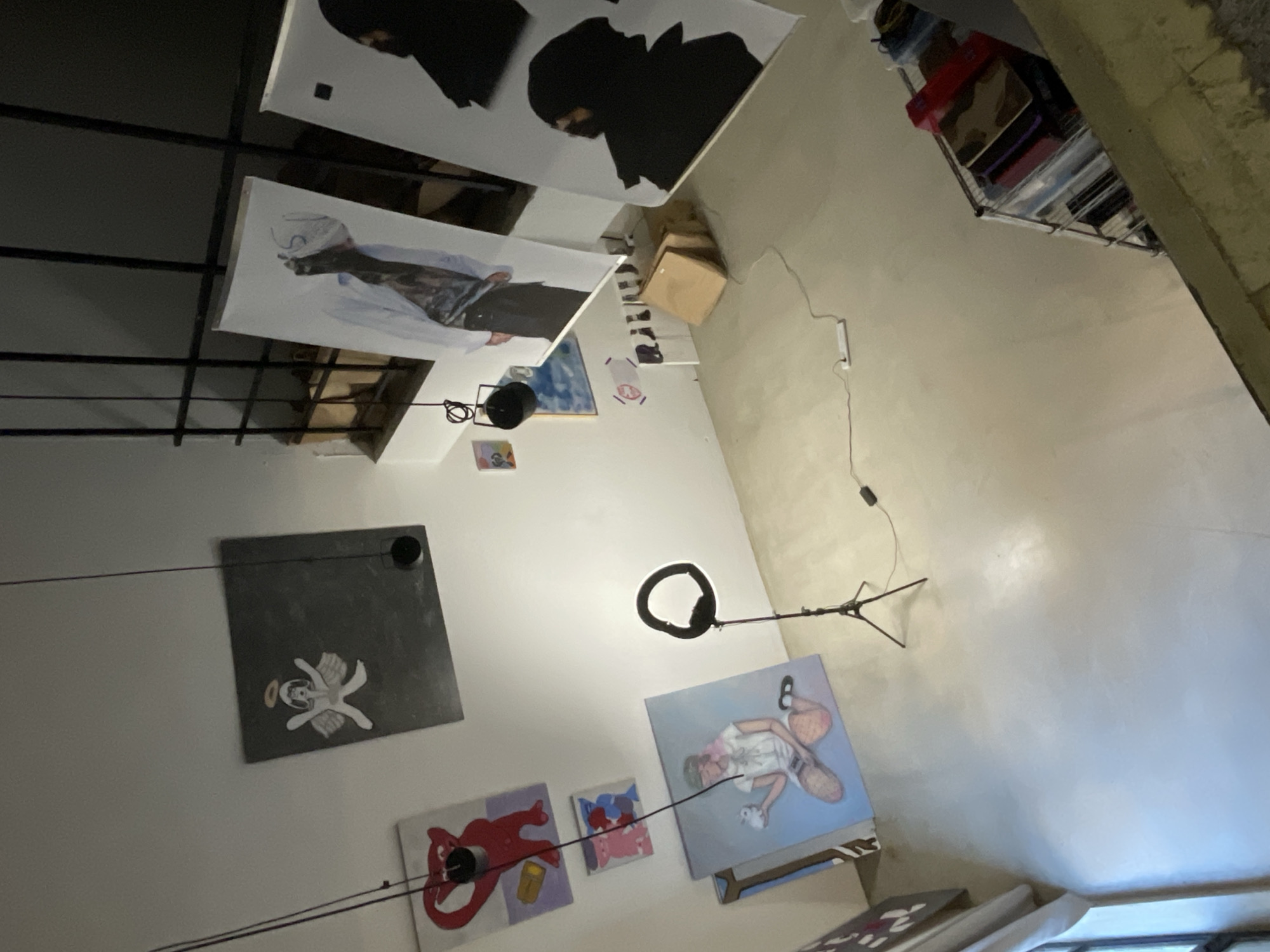 The image shows a room with white walls and a high ceiling, likely an art studio or gallery space in Buenos Aires, Argentina. A metal grid structure is affixed to the ceiling, from which several large canvases are suspended, displaying portraits and abstract figures. One canvas features a person in black headgear. Additional paintings and artworks are hung on the wall at varying heights, including a gray square with a small angel depiction, a red abstract figure, and a blue painting of a person. A ring light on a tripod stands in the middle of the room, with its power cable running along the wall. A shelf with books and other items is visible in the upper right corner of the frame. The overall environment suggests an informal exhibition or a working artist's space.