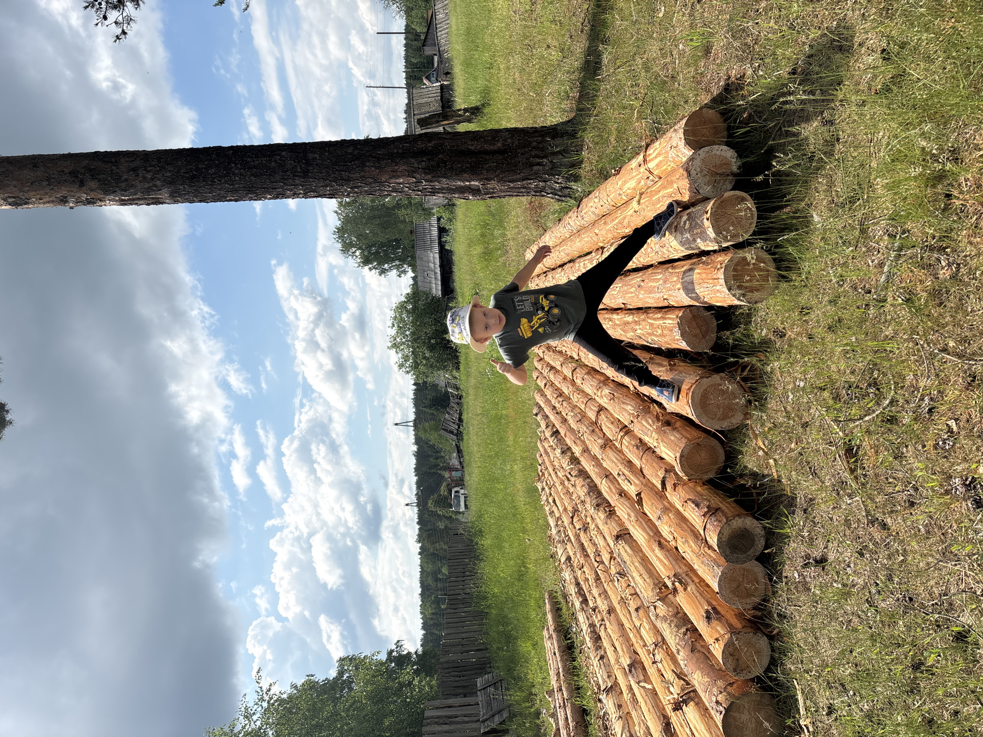 A young boy wearing a grey t-shirt with a construction vehicle graphic, black pants, and a white patterned hat is jumping with his legs spread apart in the middle of a stack of large, rough-hewn logs. The logs are laid out horizontally in rows on a grassy field. Behind the logs, a tall, dark tree trunk stands prominently on the right side of the frame. In the background, there are wooden structures, a fence, more trees, and a blue sky with white, puffy clouds. The setting appears to be rural, possibly in the city of Kurgan, Russia, given the typical landscape and wooden architecture. The boy's pose suggests an energetic and playful action.