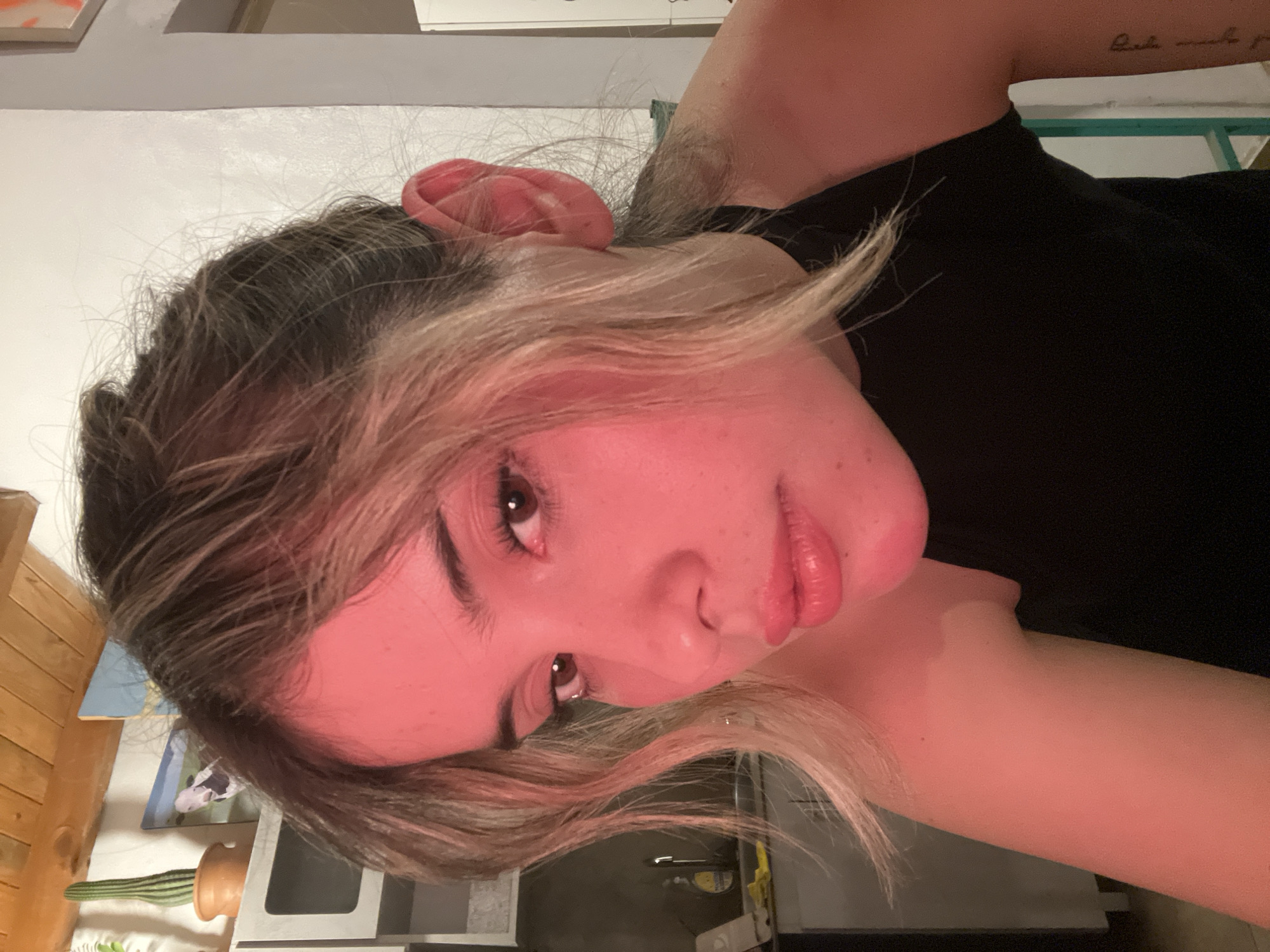 A close-up, waist-up, indoor portrait of a young woman with blonde and brown hair is captured under red lighting. She is looking directly at the camera with her mouth slightly open. She is wearing a black sleeveless top. The background includes a kitchen counter with a sink and a bottle of dish soap, a wooden ceiling, and a white wall. There is also a cactus on a shelf in the background. The image does not depict any specific location in the city of Sumalao, Argentina.