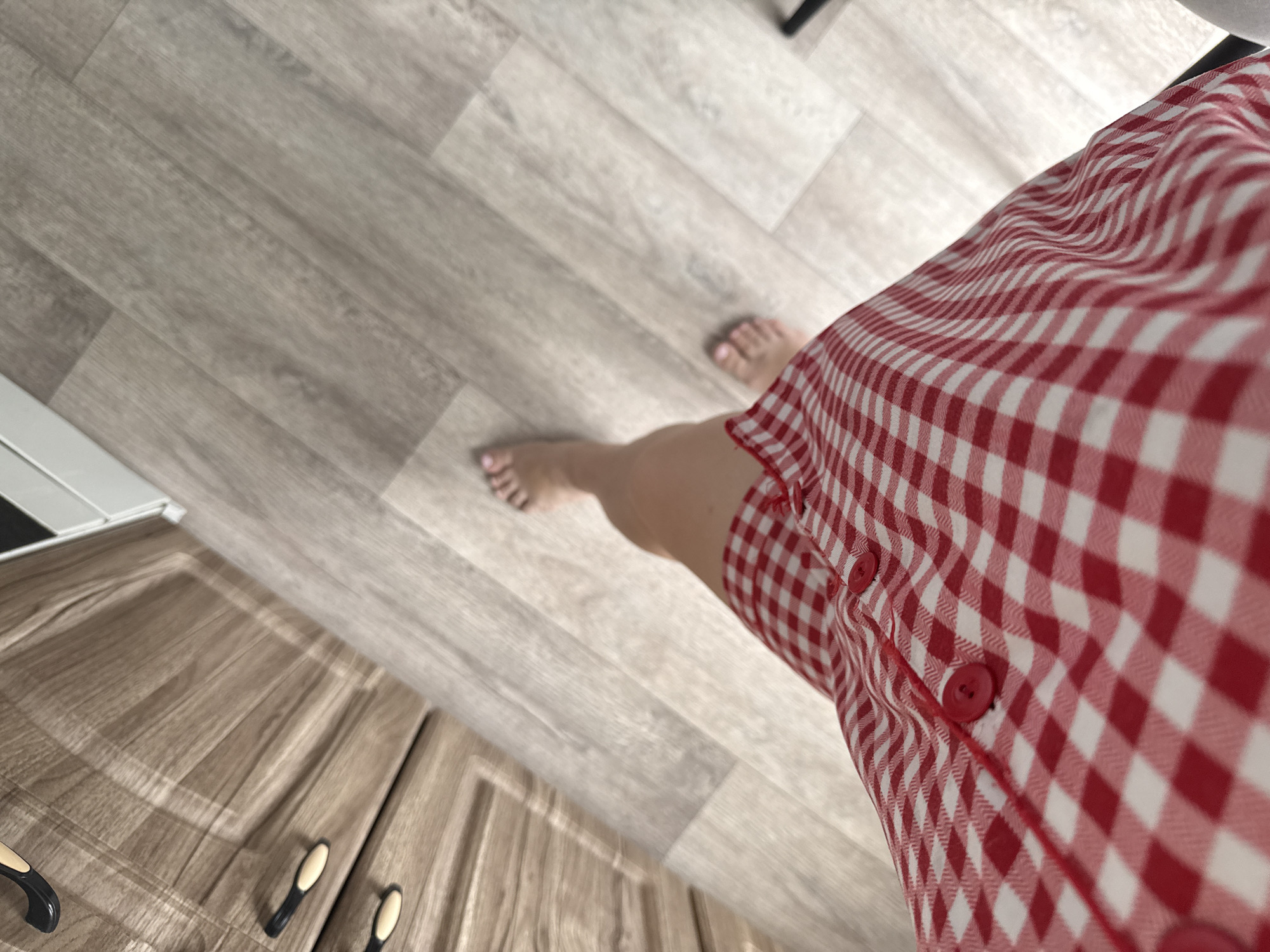 An overhead, slightly angled shot shows a person's legs from the knees down, standing on a light-colored wood-grain floor. The person is wearing a red and white gingham dress, with two red buttons visible on the front. The floor extends into a hallway or room with wooden cabinetry on the left side of the frame, featuring black and tan drawer pulls. The overall scene appears to be indoors, possibly in a residential setting. The location in Stary Oskol, Russia is not ascertainable from the visual information provided.
