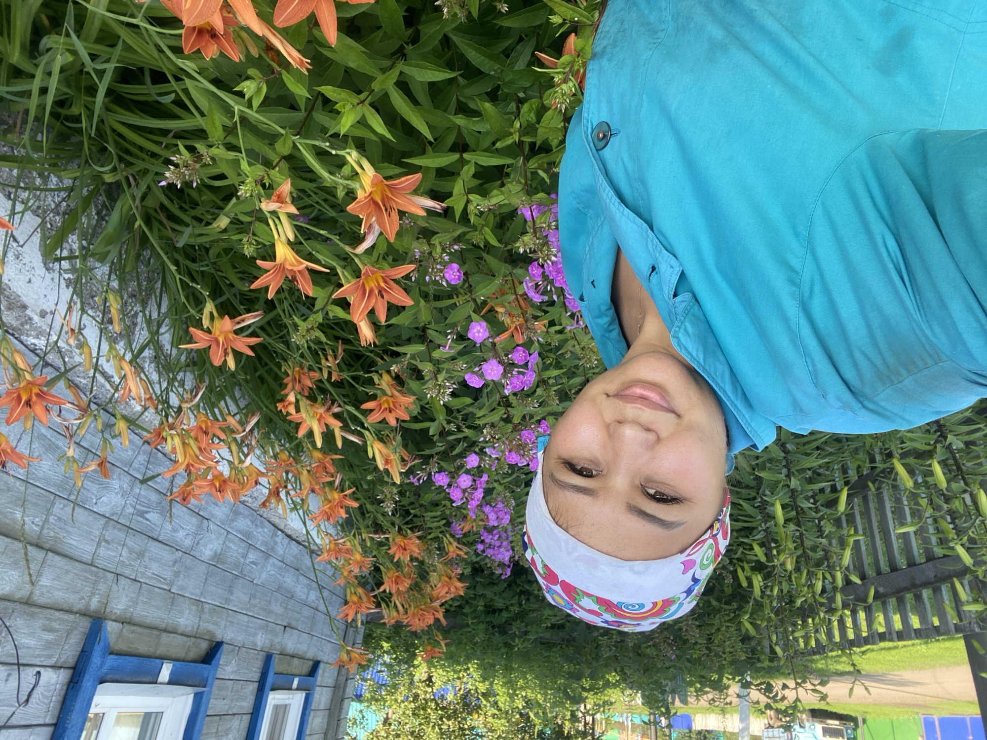 In Maloarslangulovo, Russia, a woman is taking a selfie outdoors. She is wearing a blue shirt and a colorful headscarf. She is standing in front of a flower garden with orange lilies and purple flowers. Behind the flowers is a wooden fence and a house with blue-trimmed windows. The house is made of gray wooden planks. In the background, there is greenery and a light blue truck parked on a dirt road. The lighting suggests it is daytime, possibly morning or afternoon.