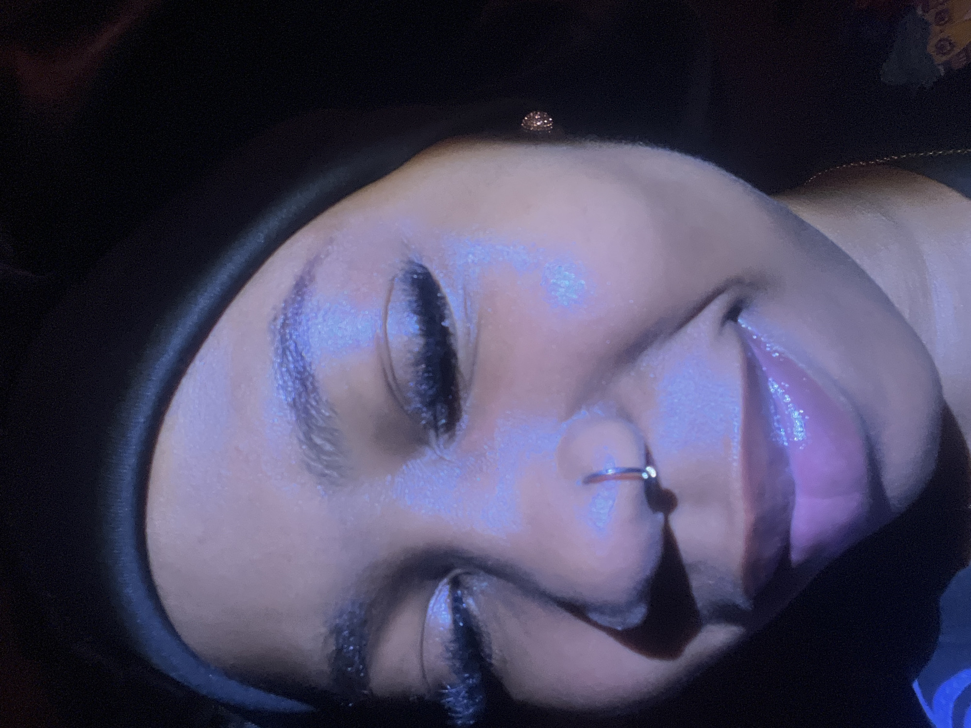 The image is a close-up, waist-up, portrait of a young Black woman wearing a black headwrap. She has on dramatic false eyelashes, a nose ring, and a shimmering pink lip gloss. The lighting is moody, with a prominent blueish cast, casting shadows and highlighting her features. Her expression is a soft smile, with her eyes partially closed, suggesting a candid or relaxed moment. There is a hint of a gold chain around her neck. The background is dark and out of focus, providing no discernible environmental context related to Rafin Par, Nigeria or any other location.