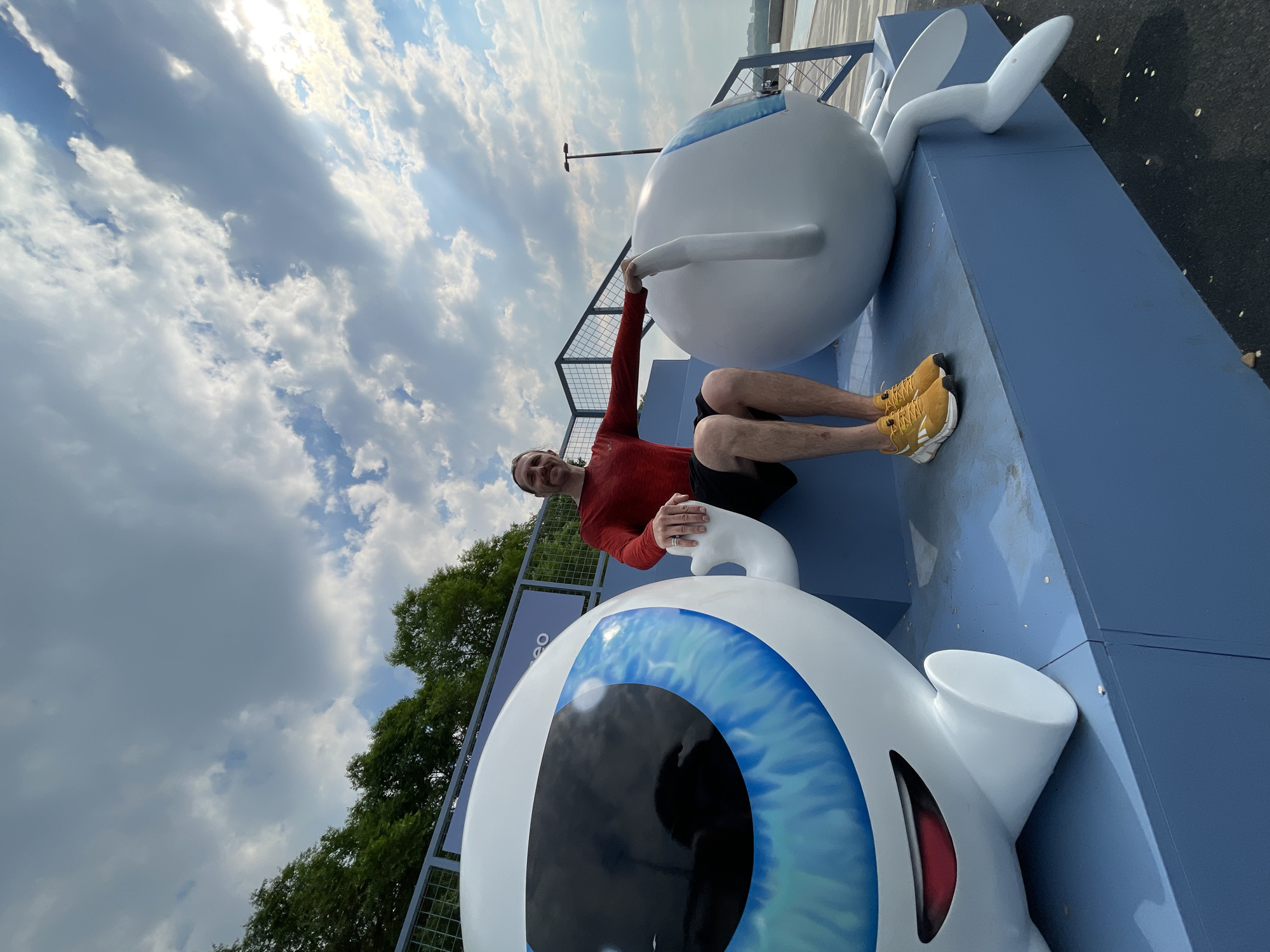 A man in a red long-sleeved shirt and black shorts is sitting on a blue tiered platform. He is smiling and looking towards the camera with his right arm extended, touching a large, white, cartoon-like eyeball sculpture to his right. To his left is another similar eyeball sculpture, facing the man. The sculptures appear to be part of an outdoor art installation or promotional display. The background shows trees, a fence, and a cloudy sky with sunlight breaking through. The scene is outdoors, likely in a public space. There is no information in the image to indicate the location is Krasnoyarsk, Russia.