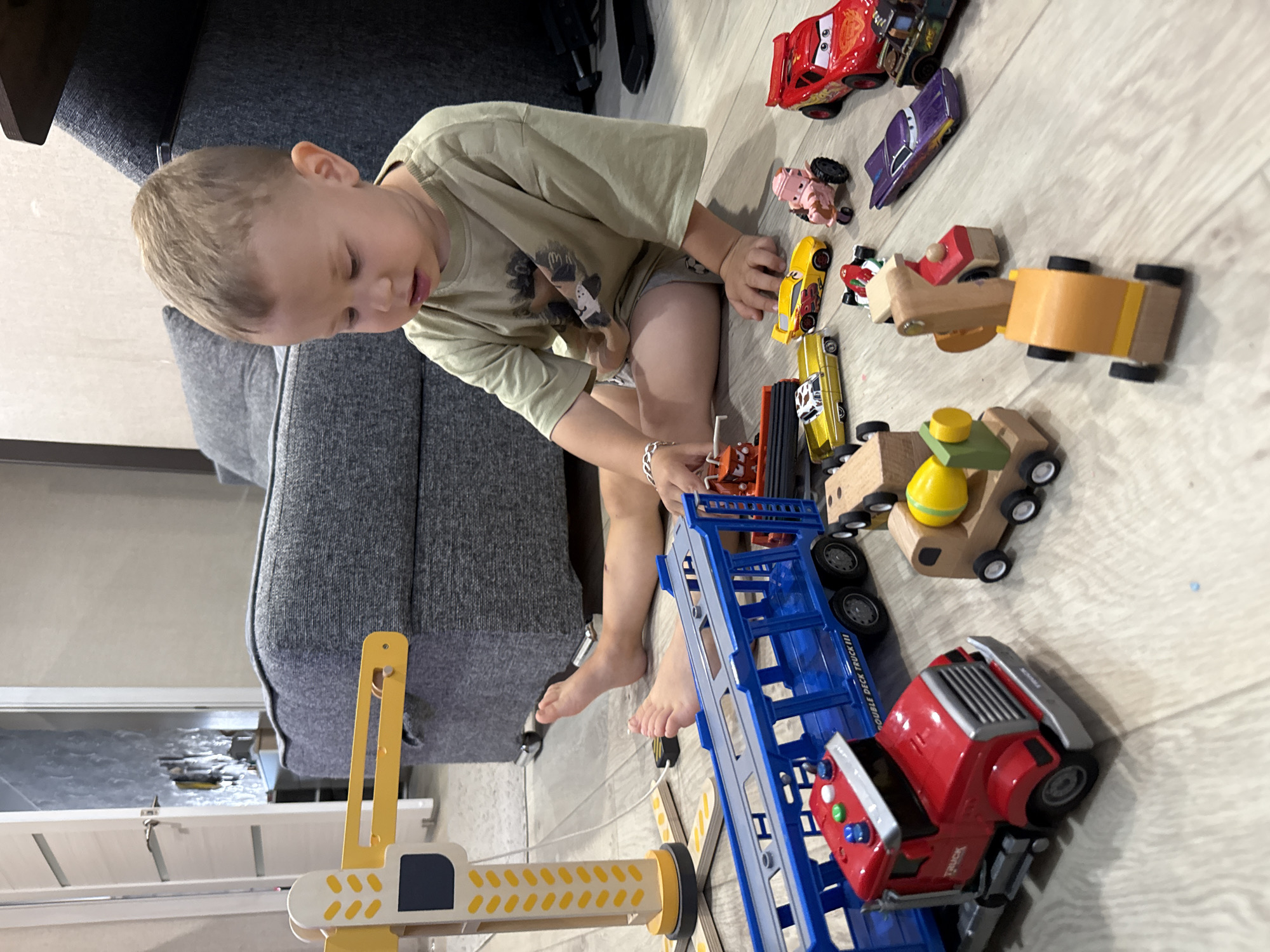 In a living room in Rusanovo, Russia, a young boy sits on the floor playing with a collection of toy vehicles. He is wearing a light brown t-shirt and gray shorts. The floor is made of light-colored wood. In front of him are various toy cars, trucks, and a construction crane. To his right, a large gray sofa is visible. The room appears to be well-lit, with natural light coming from an unseen source. The boy's attention is focused on the toys, and he appears engaged in his play.