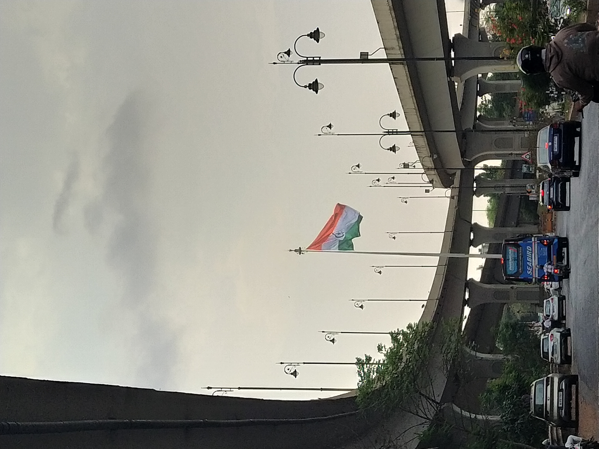 The image depicts a street scene in Panaji, India, under an overcast sky. An Indian flag is prominently displayed, flying from a tall pole amongst street lights. A flyover structure dominates the background, with vehicles including cars, buses, and auto-rickshaws visible on the road beneath it. A person wearing a helmet and jacket is positioned on a vehicle on the right side of the frame, likely a motorcycle rider. The environment includes greenery, contributing to the urban landscape.
