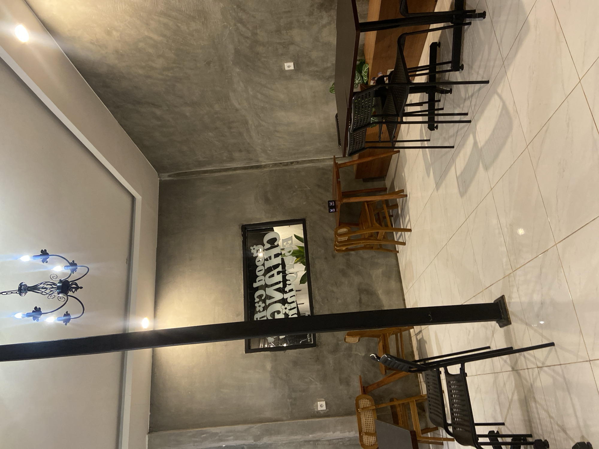 This image depicts the interior of a cafe, likely located in Sekar Mawar, Indonesia. The cafe has a modern industrial aesthetic, characterized by its gray, textured walls and a white tiled floor.
Dominating the scene is a large, black pillar that extends from floor to ceiling, suggesting structural support. To the left of the pillar, a black chandelier with five faux candles hangs from the white ceiling. Above the pillar, recessed ceiling lights provide illumination.
Several tables and chairs are arranged throughout the space. In the foreground, a black metal-framed table with two black chairs is positioned. To the left, a wooden table with a chair featuring a woven seat is visible. Further back, two wooden chairs with woven seats are placed near a wooden table. On the right side of the image, a dark brown rectangular table is accompanied by black chairs with perforated seats. Another wooden table is visible in the background on the right.
A prominent feature on the gray wall is a framed black sign with white text that appears to be a cafe logo or name, possibly reading "Good Coffee CHANO." A potted plant is visible behind the sign, adding a touch of greenery.
The overall lighting is dim, with warm tones emanating from the ceiling lights and a brighter light source on the right side of the image, casting long shadows on the floor. The environment appears to be clean and well-maintained. There are no people present in the image.