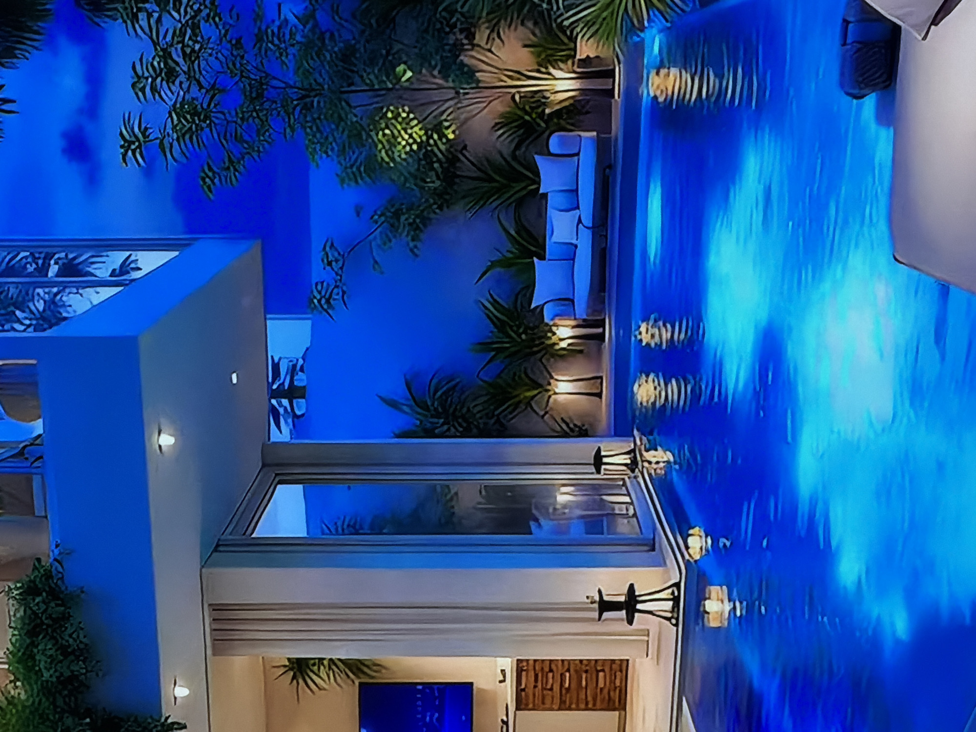 The original image, rotated 90 degrees counter-clockwise, captures a luxurious outdoor scene at night, likely a modern residential or resort property. A large swimming pool, brightly illuminated with intense blue lights, dominates the right side of the correctly oriented image. The water's surface shows vivid reflections and rippling patterns, creating a dynamic visual effect.
Adjacent to the pool stands a contemporary building with light-colored walls and large windows. Through one of these windows, an interior space is visible, featuring a flat-screen television displaying a blue image and a textured wall. Wall-mounted sconces on the building emit soft, warm light, offering a contrast to the prevalent blue.
The area is landscaped with various lush green plants and trees, some of which are highlighted by uplighting, further enhancing the dramatic atmosphere. A light-colored outdoor sectional sofa with cushions is arranged by the poolside, suggesting a comfortable seating area. Decorative lamps or lanterns are also visible near the pool's edge.
The scene is devoid of people, conveying a sense of tranquil sophistication. The dominant blue lighting, combined with strategic accent lights, creates a vibrant and almost ethereal nighttime ambiance. The setting suggests a high-end, modern design intended for relaxation and enjoyment in the evening. The location context provided is Sofiivska Borshchahivka, Ukraine.