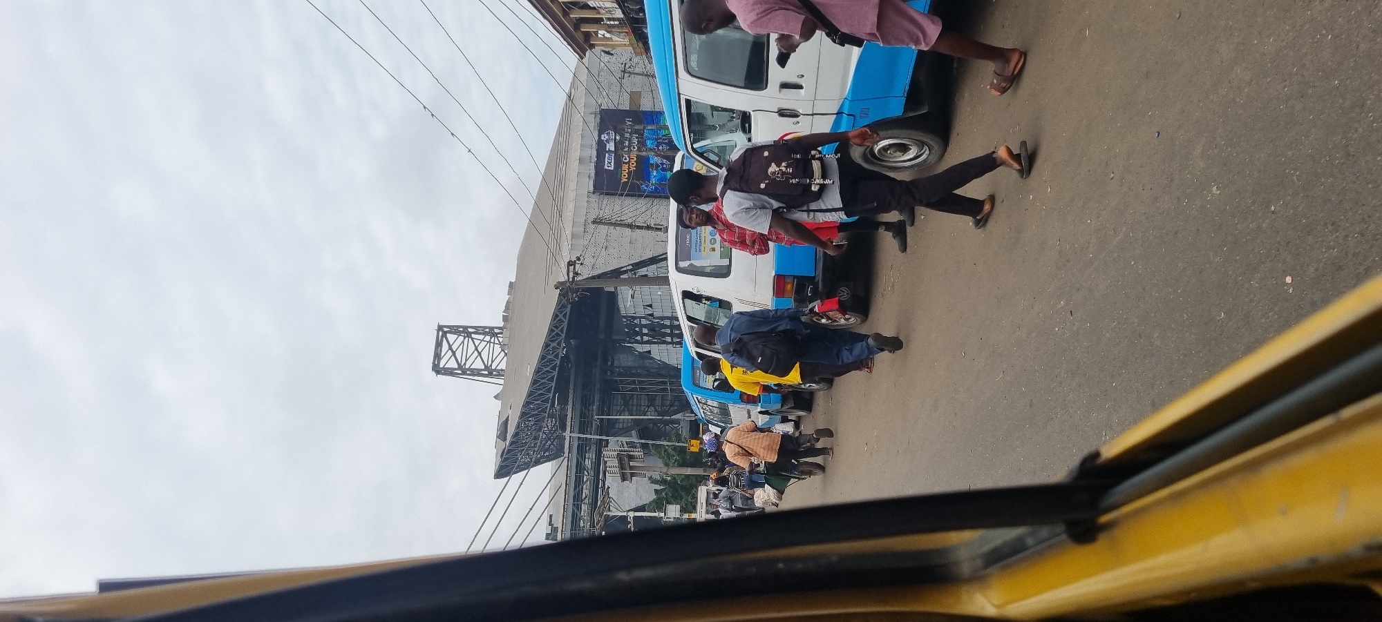 The image depicts a street scene in Ikeja, Nigeria, likely during daytime under an overcast sky. The foreground is dominated by the interior of a yellow vehicle, possibly a van or bus, with a dark frame obscuring a portion of the view. The street is paved and appears to be busy with vehicular and pedestrian traffic.
In the mid-ground, a line of blue and white minibuses, characteristic of public transportation in Nigeria, is parked or moving slowly. Several individuals are visible walking on the asphalt road. One man in a blue shirt and dark pants with a black backpack walks away from the camera. Another man in a yellow shirt and dark pants is also seen walking. A person in an orange and white striped shirt pushes a wheelbarrow. Further down the line of vehicles, a man in a red and white checkered shirt stands near a white minibus. A man in a pink shirt and light-colored shorts is seen on the right side of the image, holding a phone.
In the background, a large, modern structure with a prominent metal truss system and a sloping roof is visible. A billboard with the text "1XBET YOUR CELEBRATION! YOUR CUP!" is mounted on the side of a building. Overhead power lines crisscross the sky. The overall environment suggests an urban setting with significant activity.