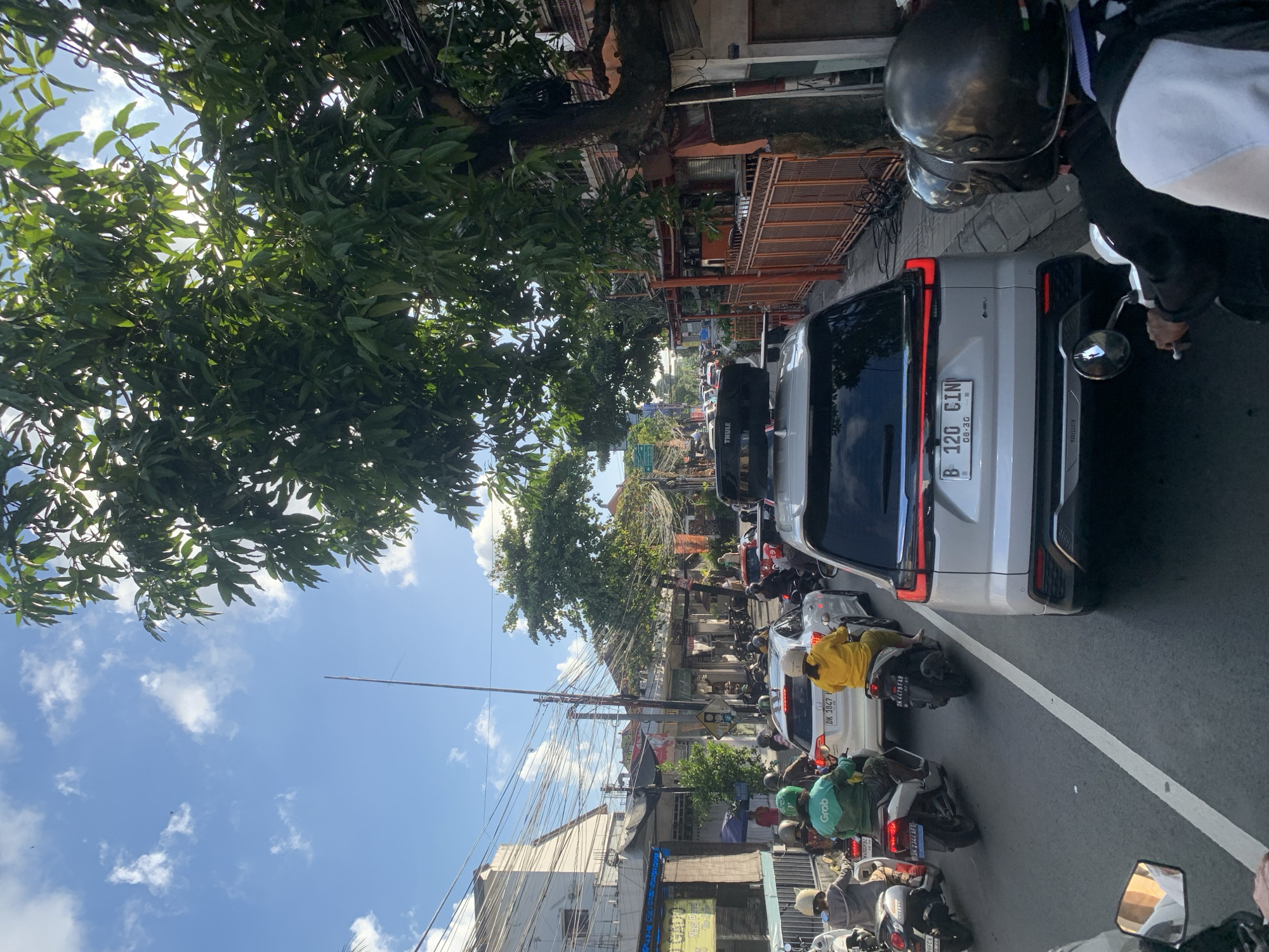 The image depicts a street scene in North Kuta, Indonesia, characterized by heavy traffic congestion. Several motorcycles, cars, and a Grab-branded vehicle are visible, all navigating a busy road lined with buildings and lush trees. The sky is partly cloudy with bright sunshine. People on motorcycles wear helmets, indicating a focus on safety. One car has a Thule cargo box mounted on its roof. The environment appears to be a typical urban street with overhead electrical wires and storefronts. The overall impression is one of a bustling day in a Southeast Asian city.