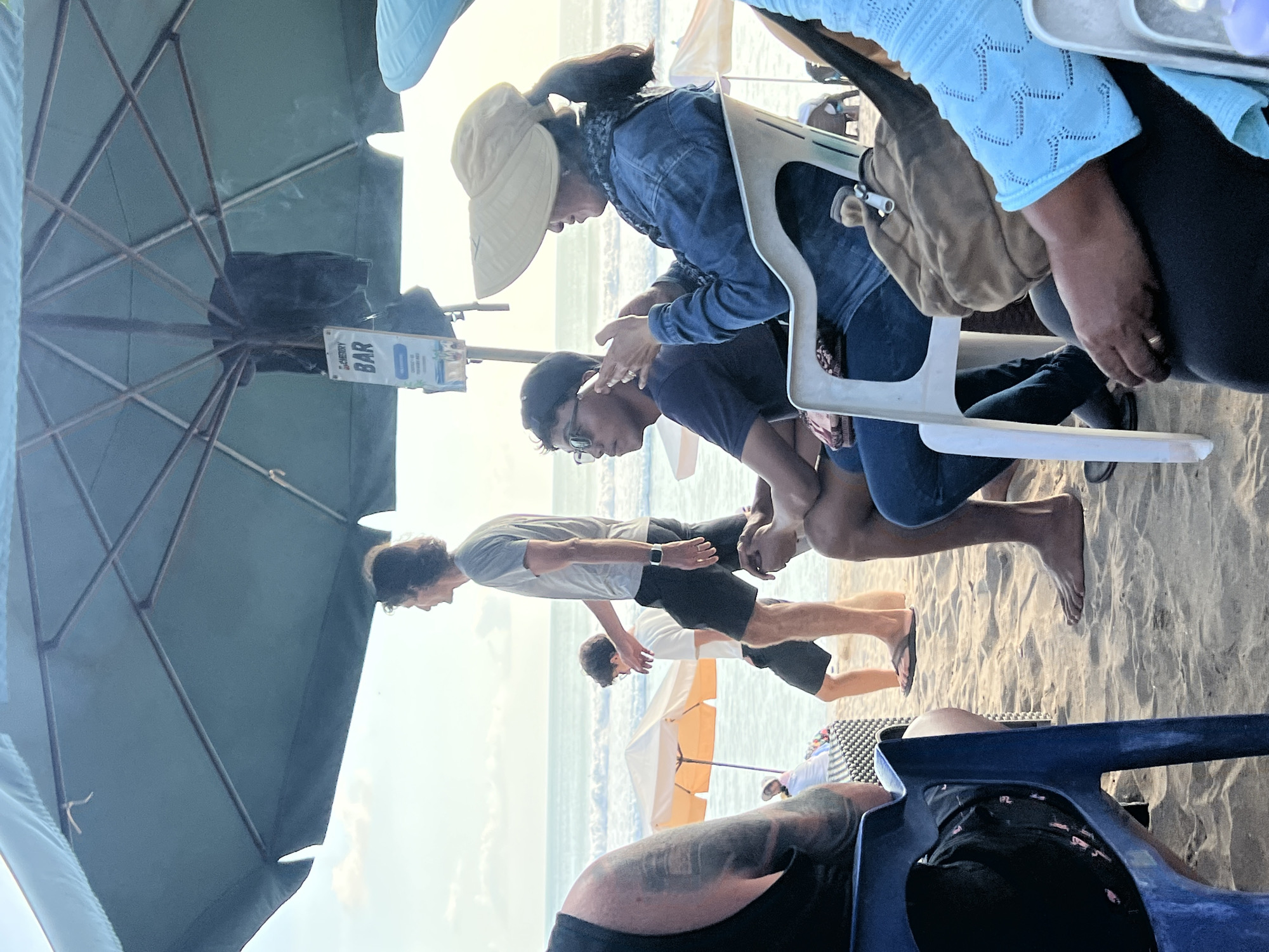 A beach scene in Kuta, Indonesia, with several people sitting and standing under a large teal umbrella. In the foreground, a person in a black tank top is seated in a blue plastic chair, facing left. To their right, a person in a denim jacket and a wide-brimmed hat is seated in a white plastic chair, holding a smartphone to their ear. Next to them, a person wearing glasses is seated in a dark shirt. In the middle ground, a man in a grey t-shirt and black shorts walks towards the right, with a man in a white t-shirt and black shorts walking in front of him. The background shows the ocean with waves and a sandy beach with other umbrellas. A sign reading "BAR" is visible on a pole under the main umbrella. The lighting suggests it is either early morning or late afternoon.