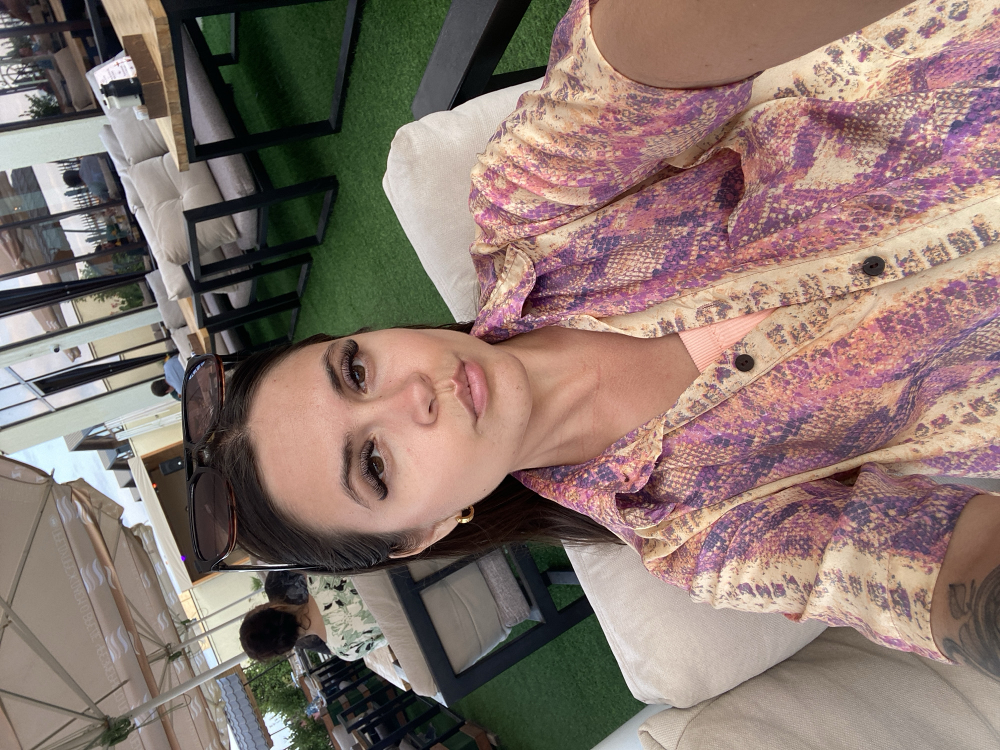 A medium shot photograph captures a woman taking a selfie in an outdoor seating area. She is positioned in the foreground, seated on a light-colored couch, and her face is the primary focus. The woman has long dark hair, and she is wearing a collared, short-sleeved shirt with a snakeskin-like pattern in shades of pink, purple, and yellow. She is also wearing a light pink top underneath. Sunglasses are perched on top of her head. Her facial expression is neutral, with her lips slightly pursed.
In the background, the scene depicts an outdoor restaurant or cafe with artificial grass flooring. There are several tables and cushioned seating areas, suggesting a modern establishment. Umbrellas are visible, providing shade over some of the seating. Reflections of people can be seen in the glass windows of a building in the background. The overall environment suggests a warm, possibly summer day. The presence of outdoor seating and a restaurant setting indicates a public space in an urban or semi-urban environment. Given the context provided, this scene is located in Derbent, Russia.
