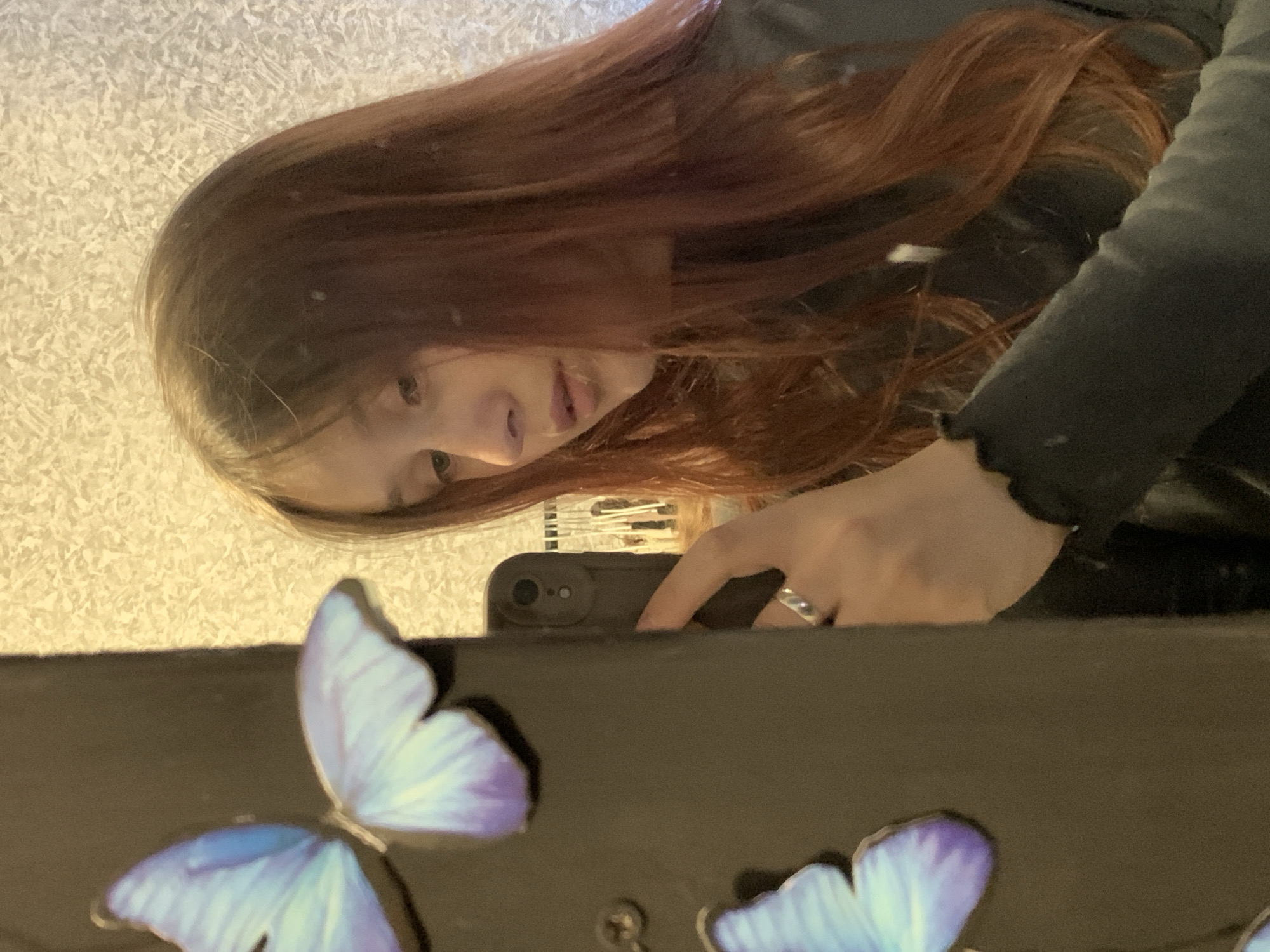 The image displays a mirror selfie of a young person with long, reddish-brown hair. Their reflection shows them partially, looking downwards, with light skin and a dark, long-sleeved top. A silver ring is visible on a finger of their right hand, which holds a smartphone in a dark case, indicating they are taking the photo.
The mirror rests on a dark surface, in front of which two decorative butterflies with shades of blue and purple are prominently displayed in the foreground. Behind the person's head, a light-colored, textured wall, possibly wallpaper, is visible, suggesting an indoor setting. The lighting appears soft, consistent with interior illumination. No specific emotion is clearly discernible from the partial view of the face. There is no visible text within the image. This scene takes place in an indoor environment, consistent with a personal space in Фоки, Russia.