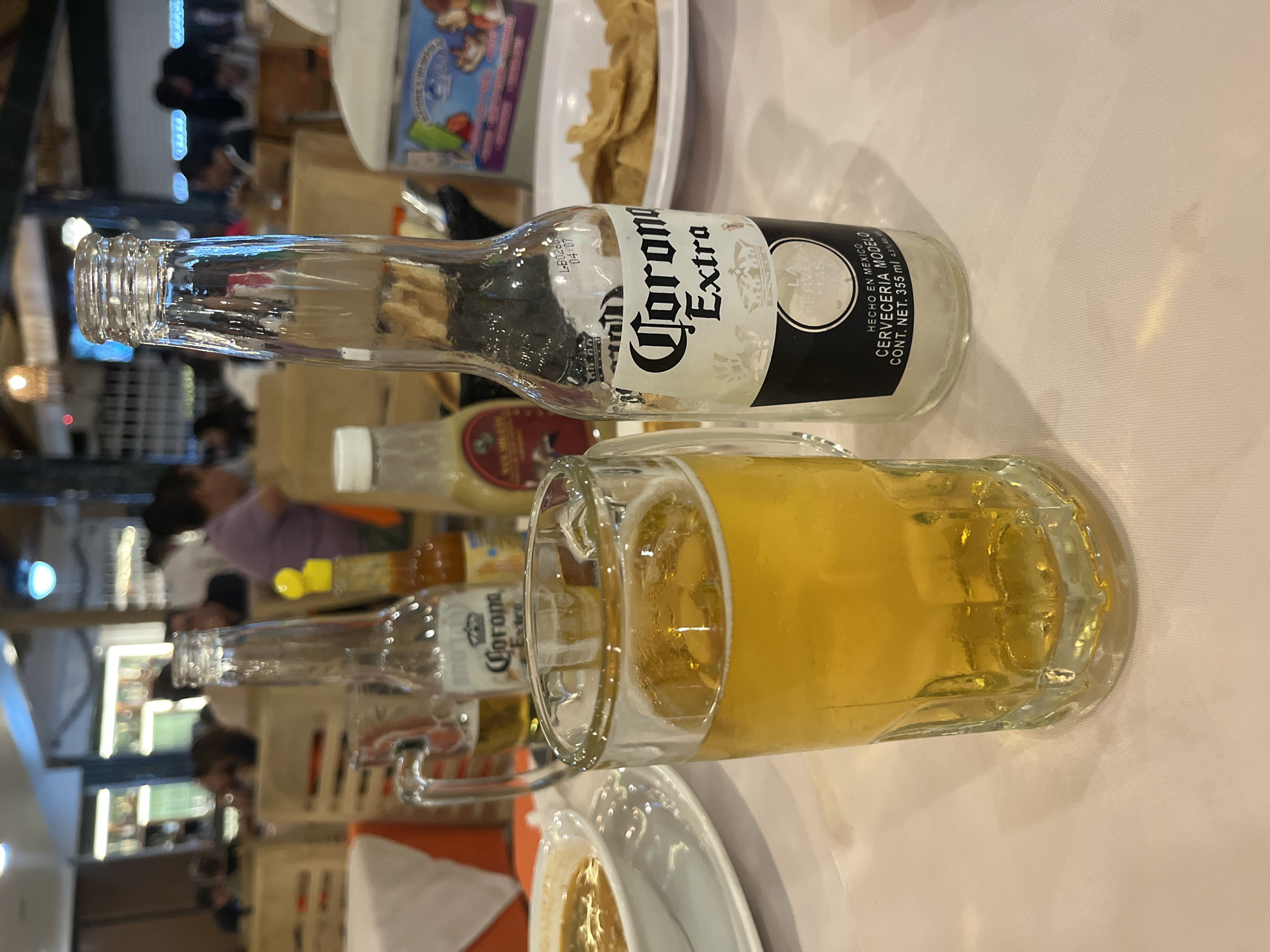 The scene depicts a table with an empty bottle of Corona Extra beer and a glass mug filled with light-colored beer. The Corona bottle is positioned horizontally on the table, with the label clearly visible. Behind it is another Corona bottle and several bottles of sauces. The beer mug is also lying horizontally and is close to an unfinished meal. The background shows a blurred view of people sitting at tables, suggesting a restaurant or bar environment. The image was captured in San Bernardino Tlaxcalancingo, Mexico.