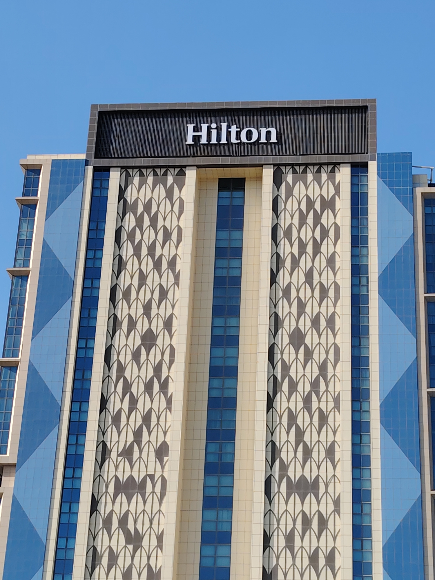 The image shows the facade of a modern hotel building in the city of Konigil, Uzbekistan. The building has a distinct architectural style with a repeating pattern of semi-circular shapes in dark grey and white on a light beige background. Large blue glass windows are interspersed with this pattern and on the side of the building. At the top of the building, a large sign displays the word "Hilton" in white lettering against a dark, textured background. The sky above is clear and blue.