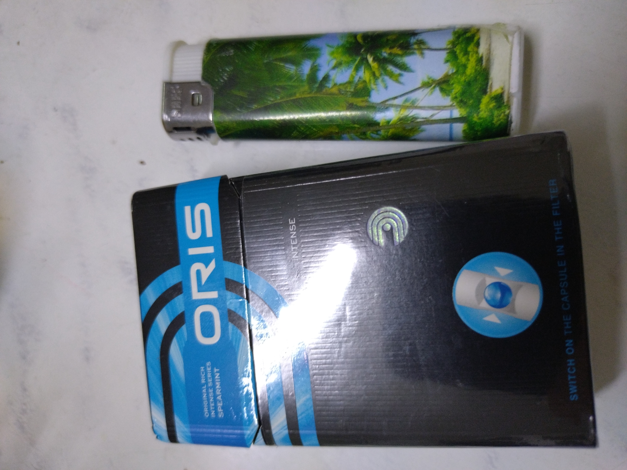 A pack of ORIS spearmint cigarettes and a disposable lighter with a tropical palm tree design are placed on a white, textured surface. The cigarette pack is primarily black with blue accents and features the ORIS logo. The lighter is green with a printed image of palm trees and a beach. The scene appears to be indoors, likely on a table or desk. No specific location in Stara Zagora, Bulgaria is identifiable from the image.