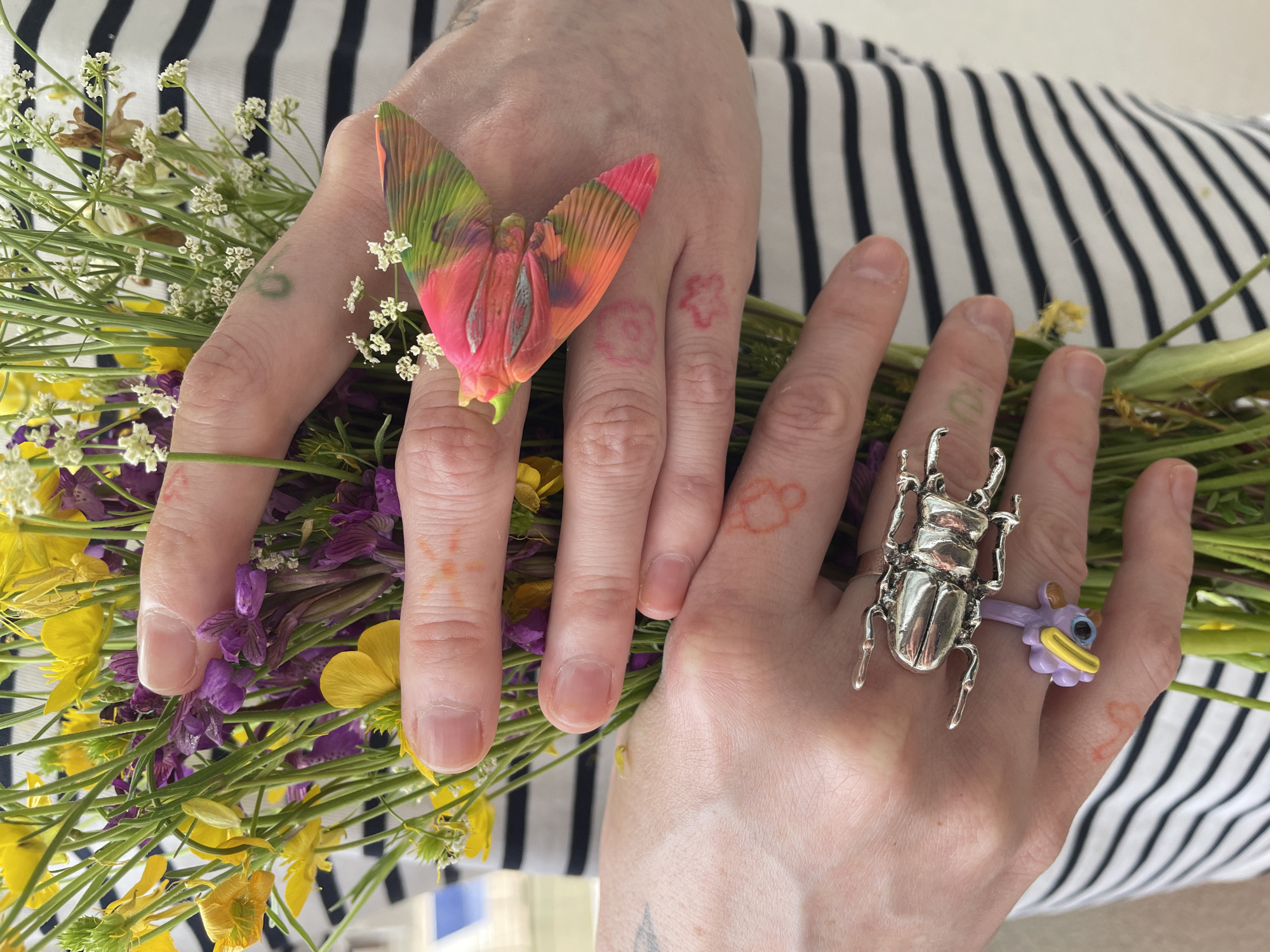 Two hands, adorned with rings and faint tattoos, hold a bouquet of wildflowers. The left hand features a ring with a colorful, petal-like design, while the right hand wears a silver beetle ring alongside a cartoonish purple and yellow ring. The background is a white and black striped fabric. There is no indication of the city of Bečići, Montenegro.