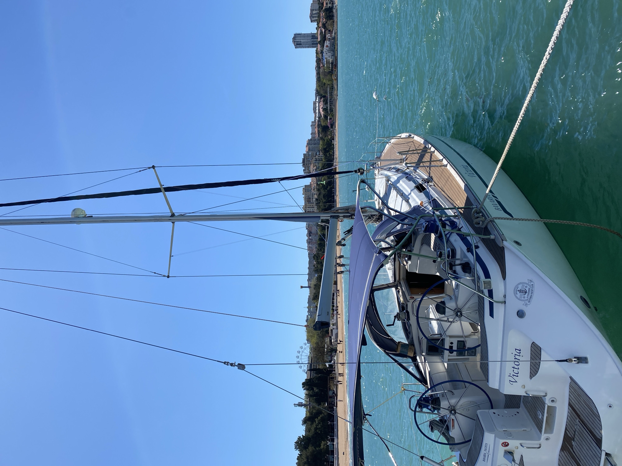 This image depicts a white sailboat, named "Victoria," moored in clear, turquoise-green water under a bright, cloudless blue sky. The sailboat is shown from its starboard side, looking towards the stern and bow. Its deck is covered in a wooden finish, and the cockpit area features two steering wheels and is partially covered by a blue canopy, likely a bimini. The name "Victoria" is prominently displayed on the stern in an elegant script, and below it, the manufacturer "BAVARIA YACHTS" is visible. A round emblem for "VICTORIA YACHT CLUB" is also affixed to the stern, along with a 'No Smoking' sign. Various lines and rigging from the mast are visible across the upper portion of the frame.
The boat is secured by a thick mooring rope extending from its side into the water. In the background, a vibrant coastline stretches along the horizon. It features a sandy beach, backed by lush green trees, and numerous buildings, including taller apartment blocks and smaller structures, suggesting an urban or resort settlement. A notable landmark on the distant coastline is a Ferris wheel, indicating a recreational area.
The scene captures a peaceful, sunny day, likely during midday or early afternoon, given the bright illumination and distinct shadows. The calm, clear water and brilliant blue sky create a serene and inviting atmosphere. The overall setting is consistent with a coastal resort town, such as Gelendzhik, Russia. No people are explicitly visible in the frame, allowing the focus to remain on the sailboat and the picturesque coastal environment.