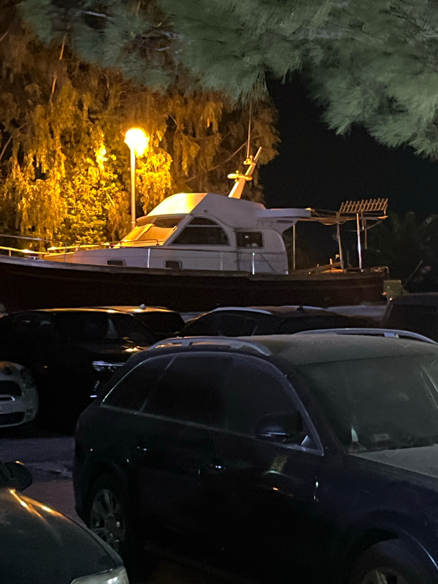 The image depicts a nighttime scene in Budva, Montenegro, featuring a boat on land, surrounded by parked cars and illuminated by a streetlamp. The boat, a substantial vessel with a white cabin and dark hull, rests on what appears to be a slipway or a designated area for storage. In the foreground, several cars are visible, including a dark-colored SUV and parts of other vehicles, indicating a parking area. Overhanging the scene are pine branches, and in the background, trees are illuminated by the warm glow of a streetlamp. The overall atmosphere is one of a quiet, possibly out-of-season, nautical setting within the urban environment of Budva.