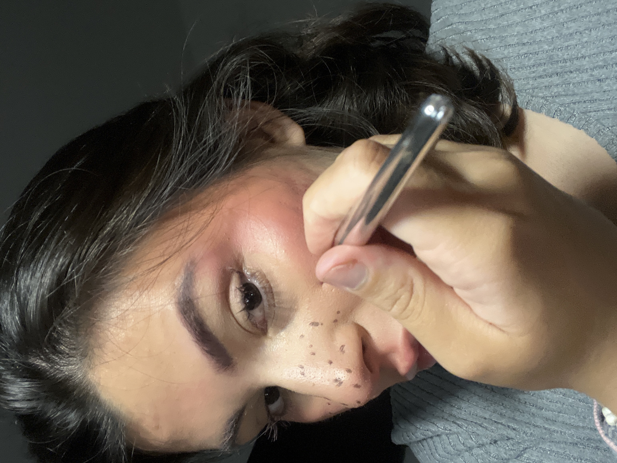 A female individual is depicted in a close-up, indoor shot, actively applying cosmetics to her face. Her left facial profile is visible, featuring dark hair, defined eyebrows, and dark eyes. Numerous small, dark-brown, irregularly shaped dots, consistent with artificial freckles, are present on her nose and the upper portion of her left cheek. A subtle reddish tone is visible on her cheek. Her right hand, with manicured fingernails, holds a slender, metallic, silver-colored applicator, which is positioned near her nose, suggesting the application or touch-up of the aforementioned freckles. She is wearing a grey, ribbed garment. The background is dark and indistinct, indicating low light conditions. The location is Kiryat Bialik, Israel.