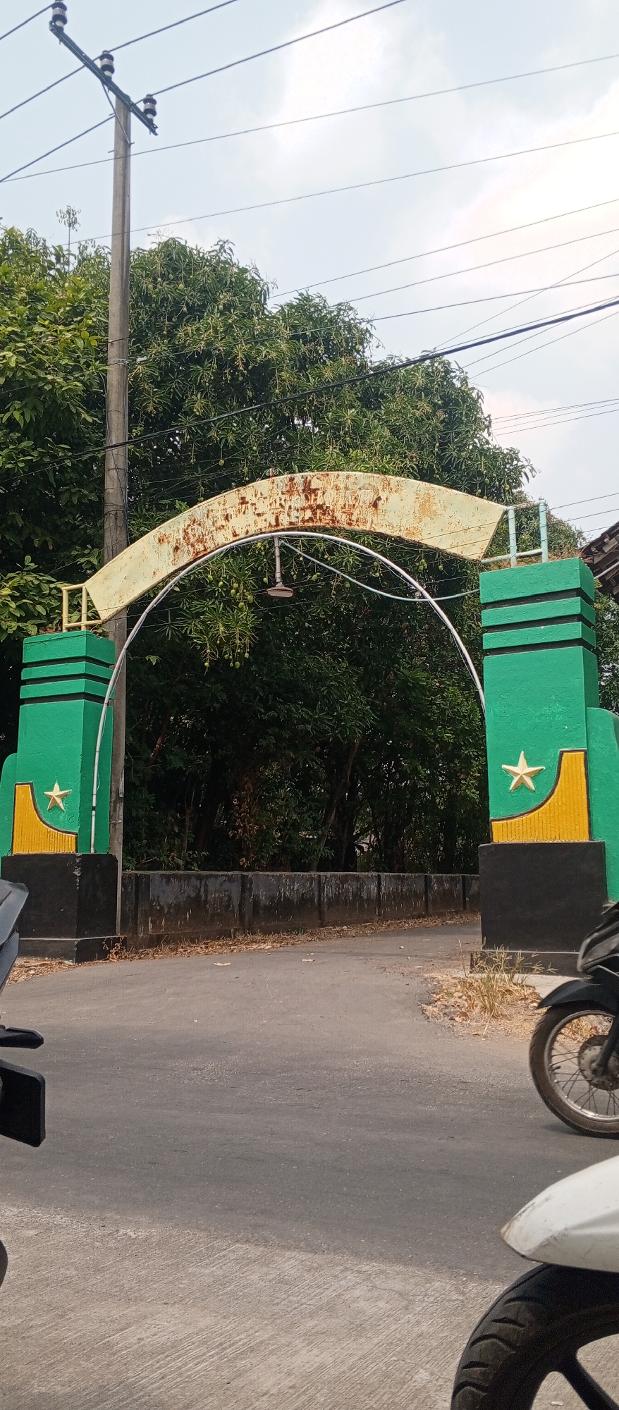 This image captures an arched gateway, likely an entrance to a residential or administrative area, situated on a road in Gulun, Indonesia. The gateway features two sturdy, green pillars adorned with yellow accents and a star insignia. A rusty, yellow-colored archway spans across the top, supporting a metal rod with a light fixture. Beyond the gateway, a dense canopy of green trees suggests a lush environment, with a clear sky visible above. The foreground of the image shows a paved road with motorcycles, one white and one black, partially visible, indicating active use of the area. The scene is captured during daylight hours, under a bright sky.