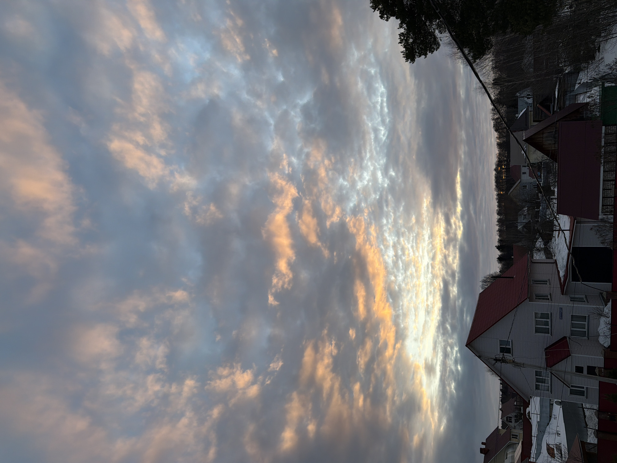 This image depicts an exterior view of a residential area in the city of Chaikovsky, Russia, specifically located at Chaikovsky Boulevard 35, as indicated by a street sign on the prominent house. The sky occupies the upper two-thirds of the frame, filled with stratocumulus clouds displaying a spectrum of colors from deep grey and pale blue to vibrant orange and pink, with the brightest light concentrated towards the lower-center horizon, consistent with either sunrise or sunset.
In the mid-ground, a two-story house with white horizontal siding and a steep, red gable roof is centrally positioned on the left. A smaller, single-story extension with a matching red roof is attached to its front facade. Multiple windows are visible on the house. A utility pole with several electrical wires runs vertically in front of this residence. The surrounding ground and rooftops of adjacent houses exhibit patches of snow, indicating a winter or early spring season. Various other residential structures with different roof colors (red, dark grey) are visible in the background and mid-ground. Bare deciduous trees and some dark evergreen foliage are present, with a dense tree line forming the distant horizon. Overhead utility wires are visible traversing the upper right portion of the scene. No individuals, actions, or specific interactions are depicted.