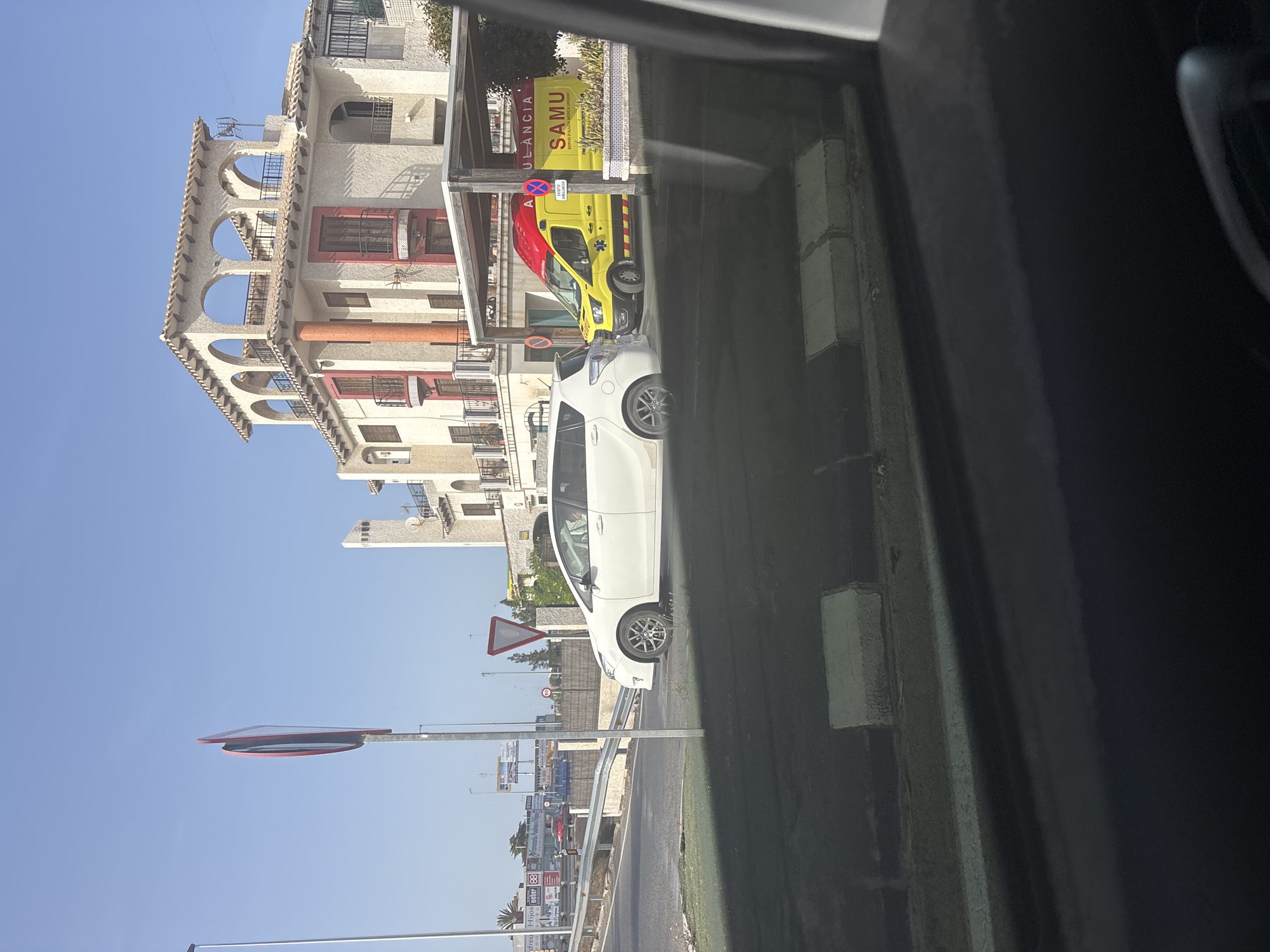 A white car is parked on the right side of a paved area in front of a building. Next to the white car, a yellow and red ambulance is parked under a covered area. The ambulance has "SAMU" written on its side. The building behind them is a multi-story structure with balconies and arched windows. In the background, there are other buildings, street signs, and a clear blue sky. The foreground shows a dark, reflective surface, possibly a car window.