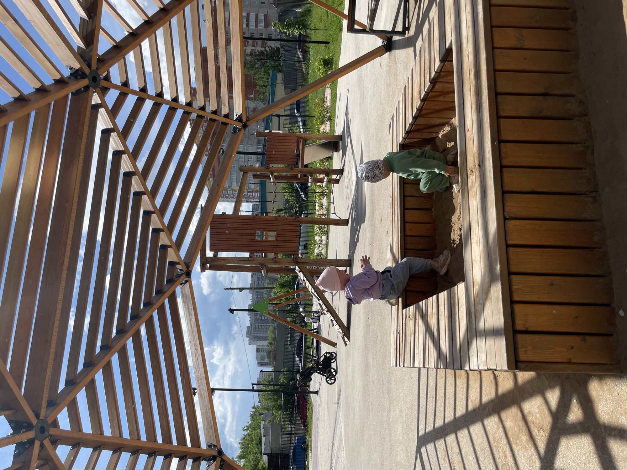 Two young children are playing in a wooden sandbox at a playground in Izhevsk, Russia, under a partially cloudy blue sky. The child on the left, wearing a pink beanie, purple hoodie, and light jeans, is seated on the raised wooden edge of the sandbox, looking towards the right. The child on the right, dressed in a green sweatsuit and a grey patterned beanie, is kneeling within the sandbox, facing left and interacting with the sand. A prominent wooden pergola with radial supporting beams and horizontal slats covers the area, casting distinct linear shadows onto the concrete ground. In the mid-ground, a black baby stroller is parked near other playground equipment, including a wooden slide and a climbing structure with green accents. Beyond the playground's paved surface, a dark fence separates the area from parked cars and distant multi-story residential buildings. Trees and green vegetation are visible behind the fence.
