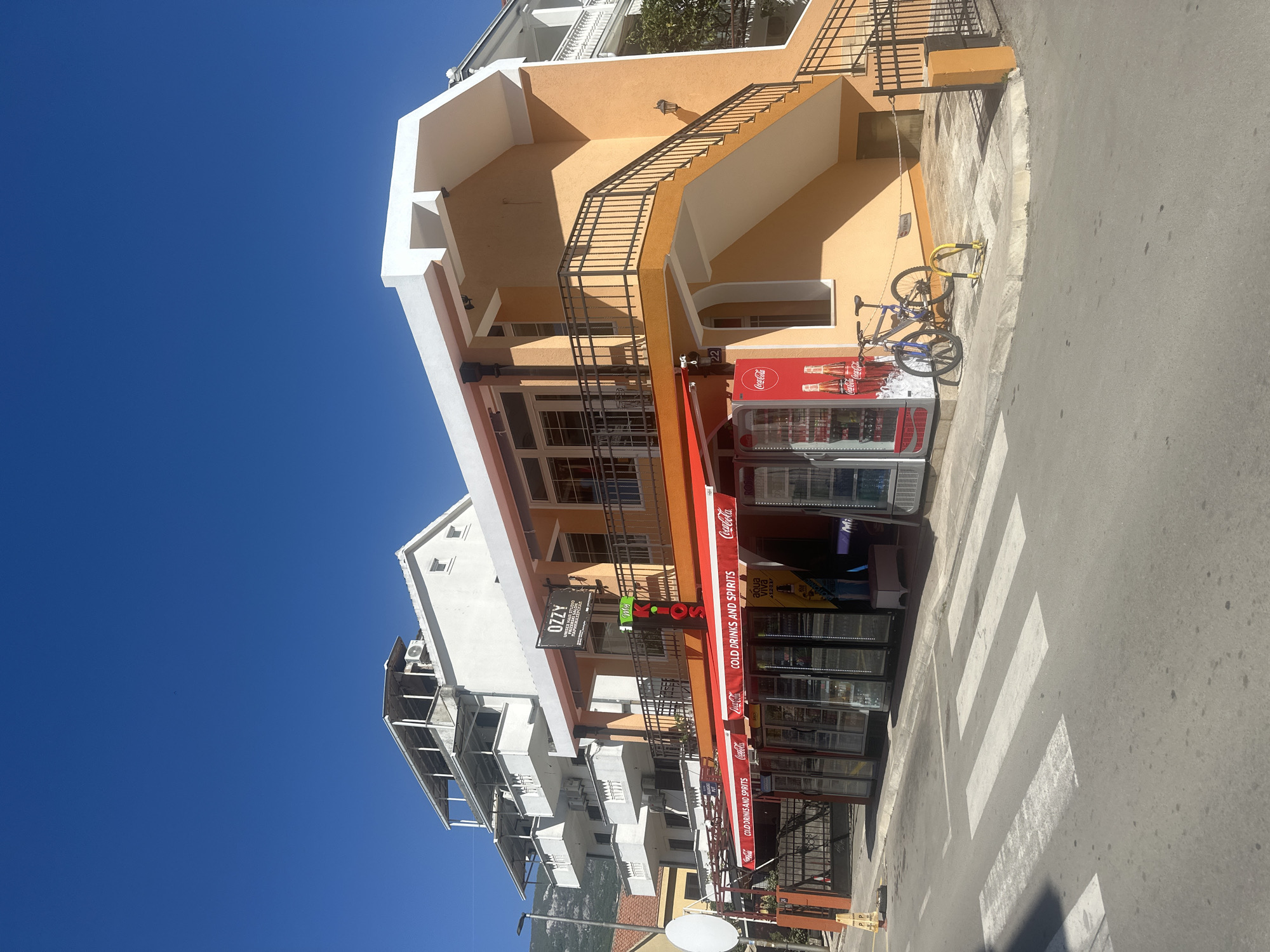 The image captures a bright, sunny street scene in Budva, Montenegro, dominated by a clear, deep blue sky. The scene features a multi-story building complex, with an orange-colored structure central to the composition. This building has an external, black metal staircase leading to upper floors and houses a ground-level shop.
The shop is characterized by a red awning extending over its entrance, displaying branding for "Coca-Cola" and text that reads "COLD DRINKS AND SPIRITS." Several Coca-Cola branded display refrigerators are visible outside the shop, containing beverages like "aqua viva" and "SPORT," with another sign potentially indicating "Mojito." Above the shop, on the orange building, a sign for "OZZY UNISEX HAIR STUDIO" is mounted, along with the number "22."
Adjacent to the orange building, to the left, is a white residential building with multiple balconies and air conditioning units visible on its facade. In the foreground, a blue bicycle is parked next to a yellow bike rack on the sidewalk. The pavement transitions into a paved street with a prominent white zebra crossing. The bright daylight casts distinct shadows from the buildings and objects, indicating a sunny day. No people are visible in the image.